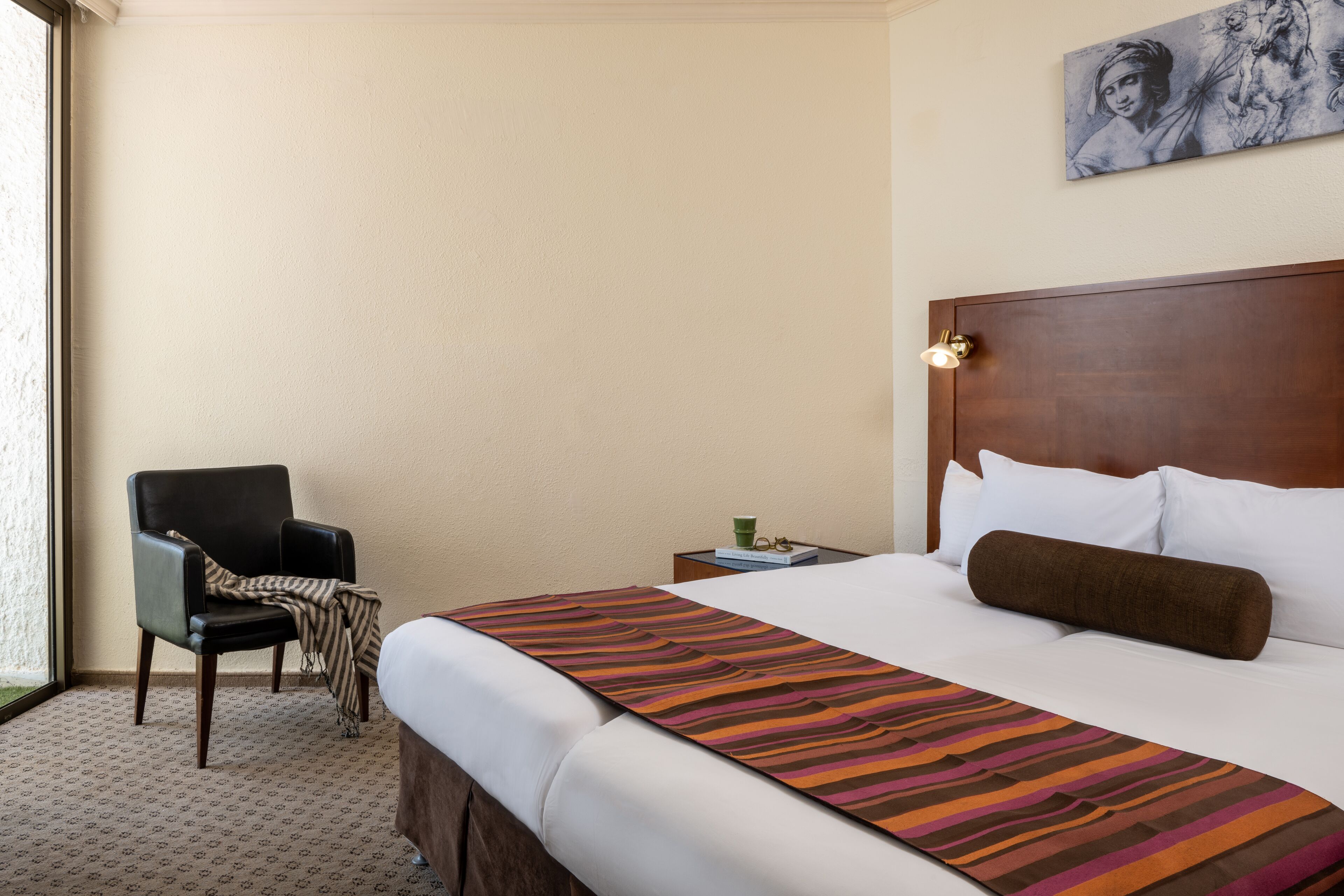 A hotel room with a bed, a chair, a nightstand, and a painting on the wall.