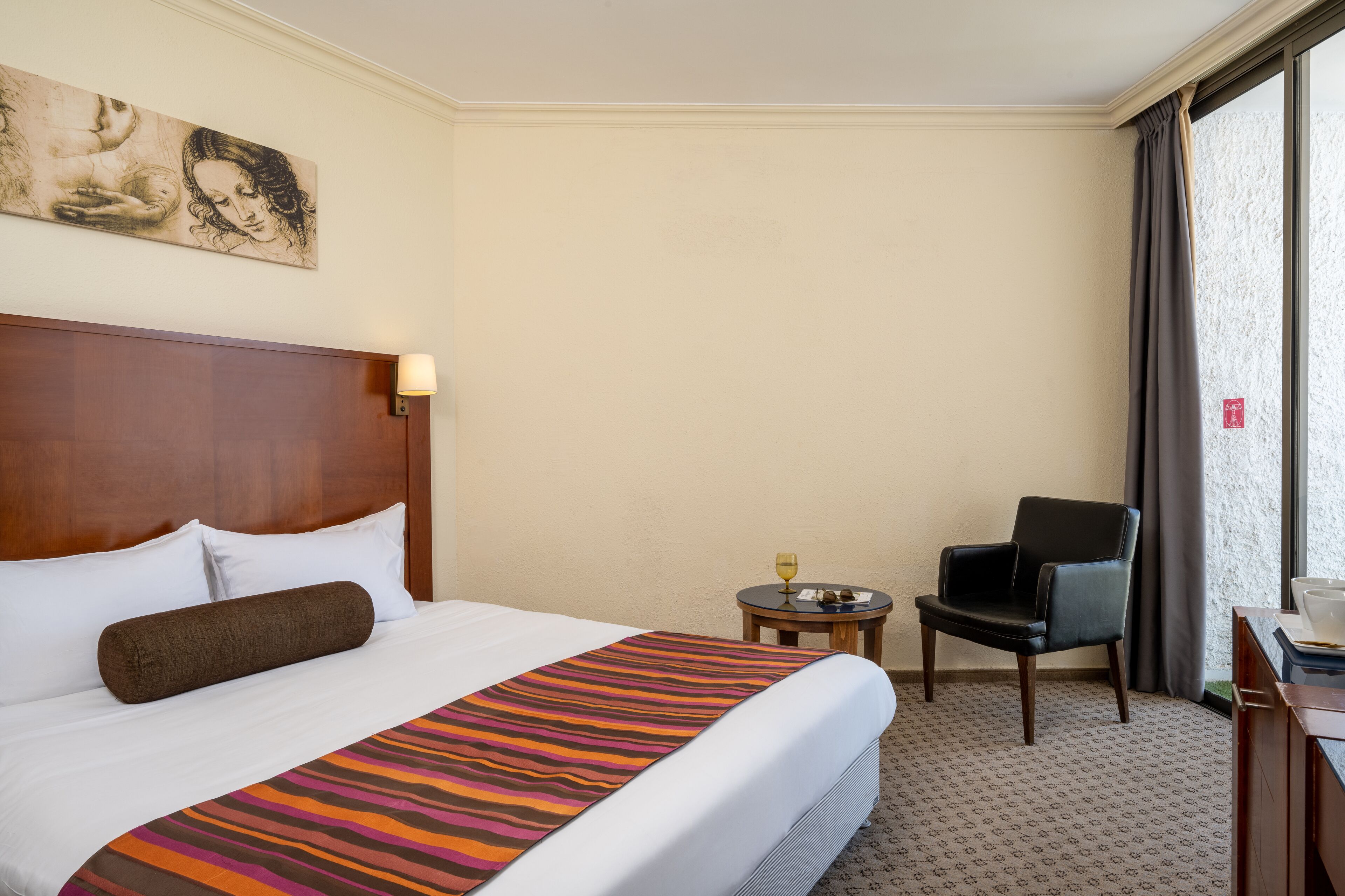 A hotel room with a large bed, a wooden headboard, a small round table with a glass of wine, a black chair, and a painting on the wall.
