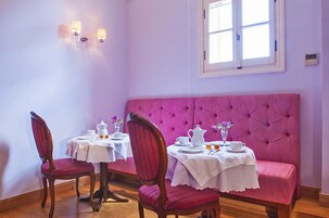 Un comedor con dos mesas, cada una con mantel blanco, cubiertos, sillas y un banco tapizado en rosa.