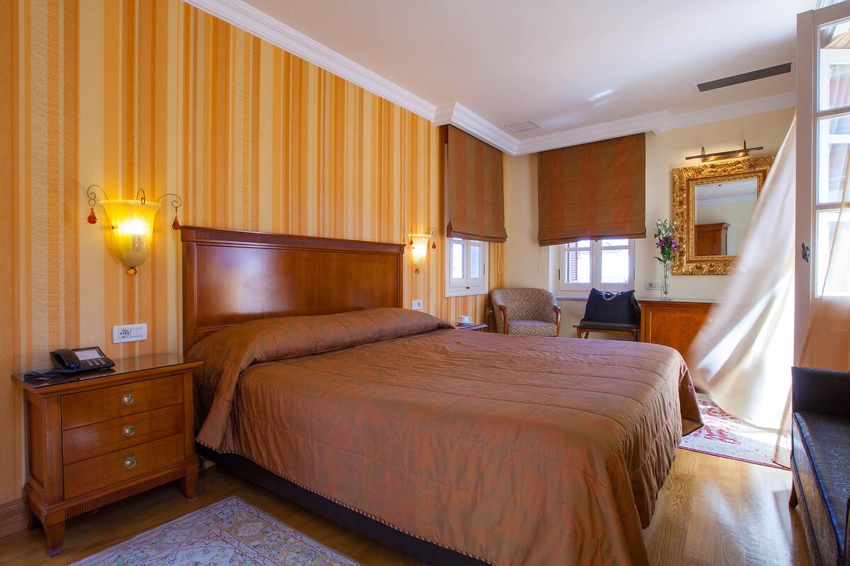 A hotel room with a large bed, a nightstand with a phone, a chair, a desk with a lamp, and a mirror.