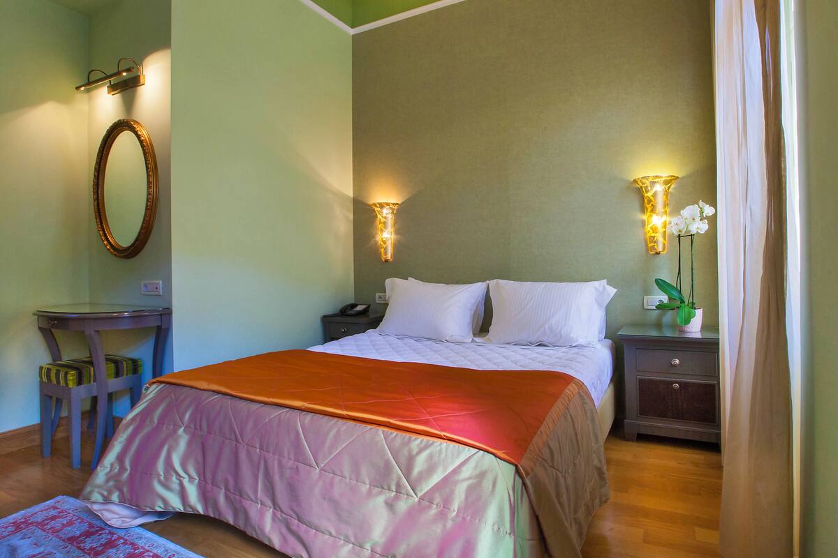 A neatly made bed with a pink and orange bedspread, a bedside table with a lamp, and a nightstand with a plant.