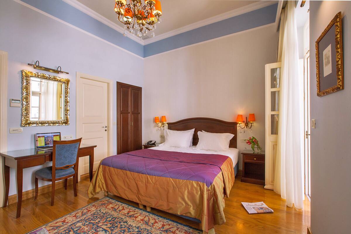 A bedroom with a bed, a desk, a chair, a mirror, and a chandelier.