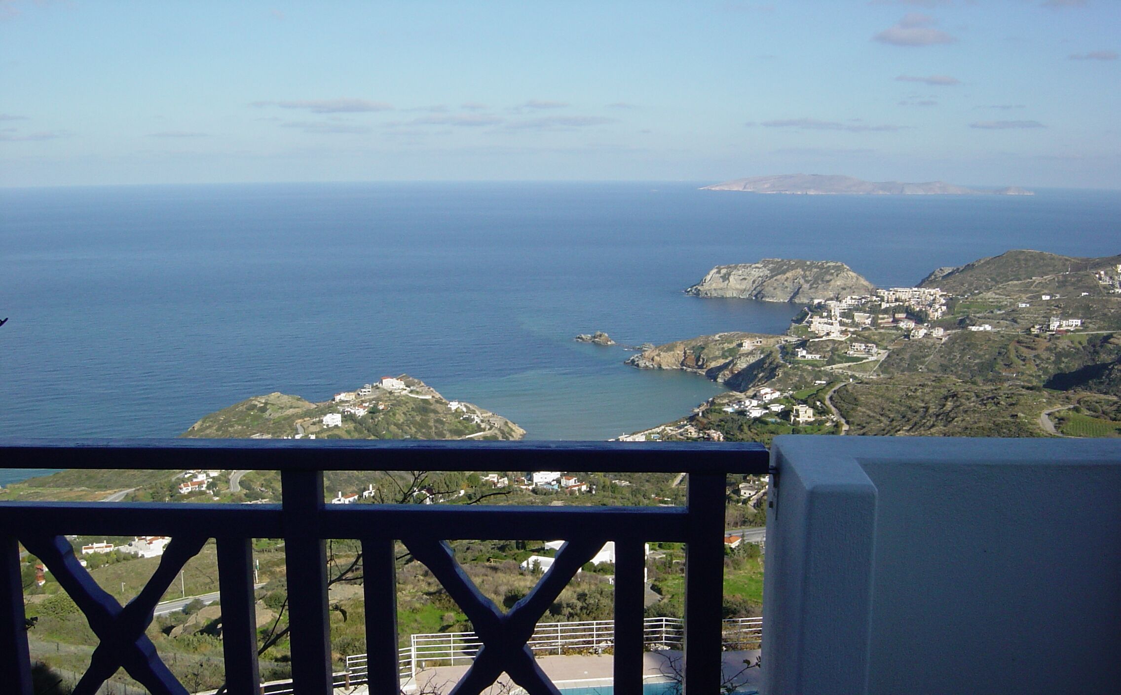A coastal view from a balcony with a railing, overlooking a sea, rocky terrain, and scattered buildings.