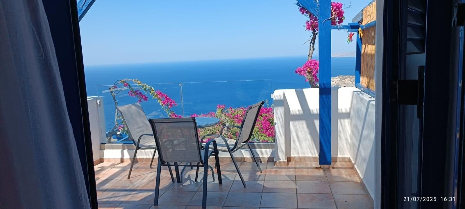 A balcony with a view of the sea, a table with chairs, and pink flowers.