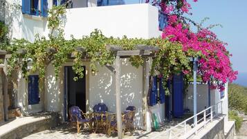 Ein weißes Gebäude mit blauen Fensterläden, einer terrassenförmig angelegten Pflasterterrasse und einer mit Grünpflanzen und rosa Bougainvillea bewachsenen Pergola.