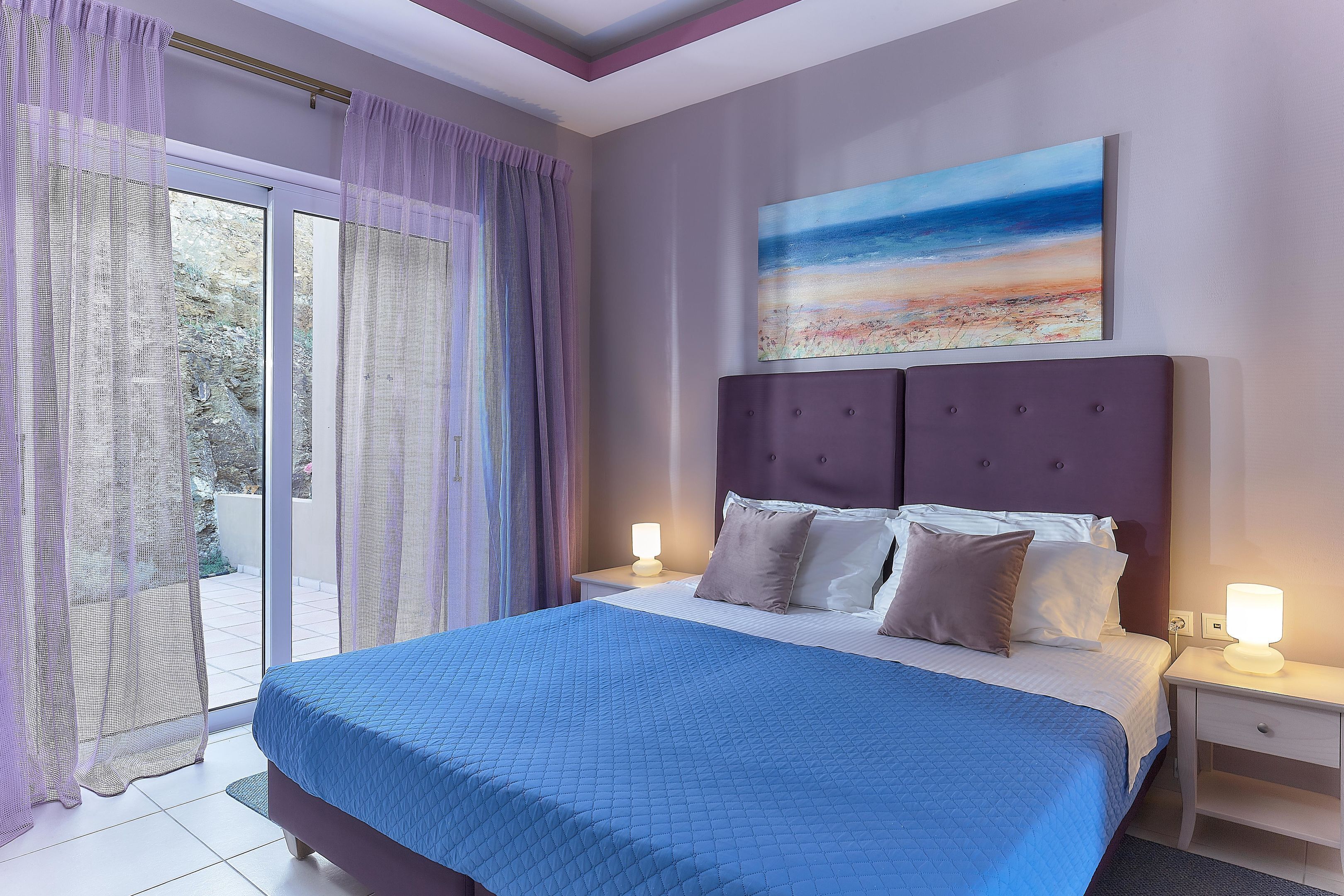 A bedroom with a large bed, a nightstand, a wall-mounted painting, and a sliding glass door with curtains.