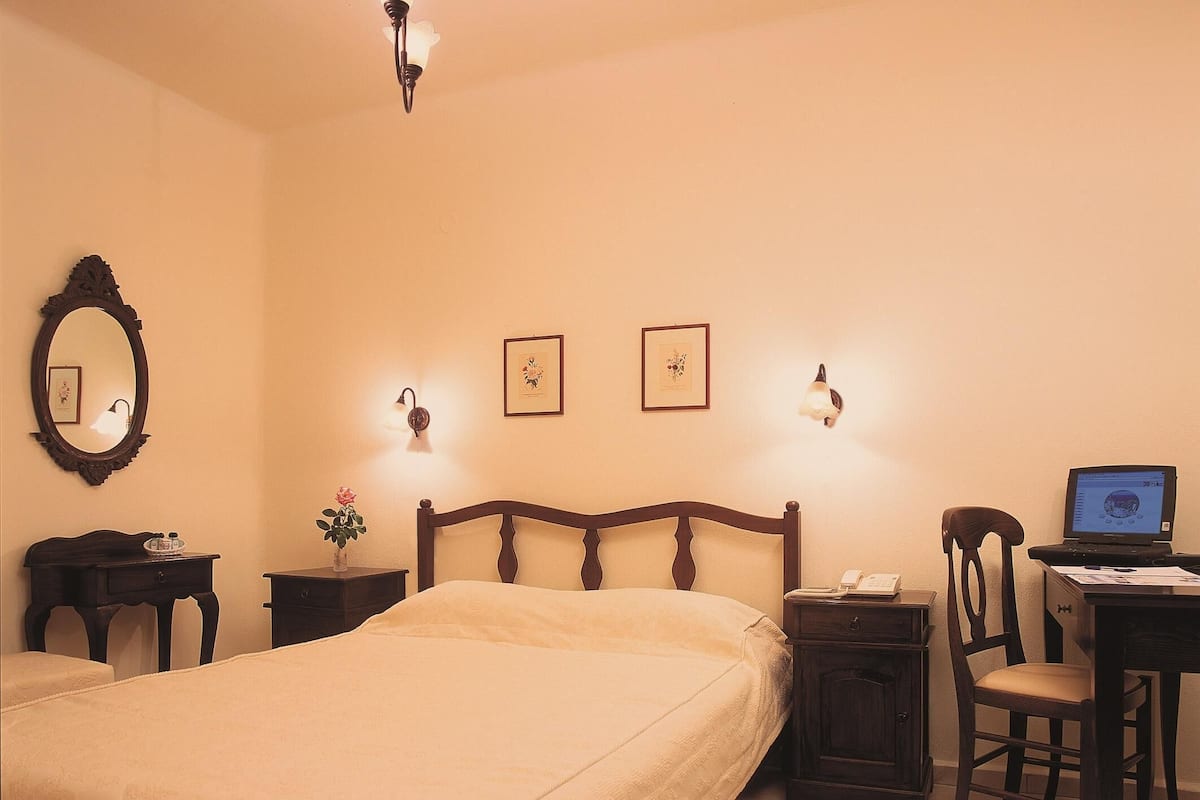 Une chambre d’hôtel avec un grand lit, un bureau avec un ordinateur, une chaise, une petite table avec un vase de fleurs et deux lampes fixées au mur.