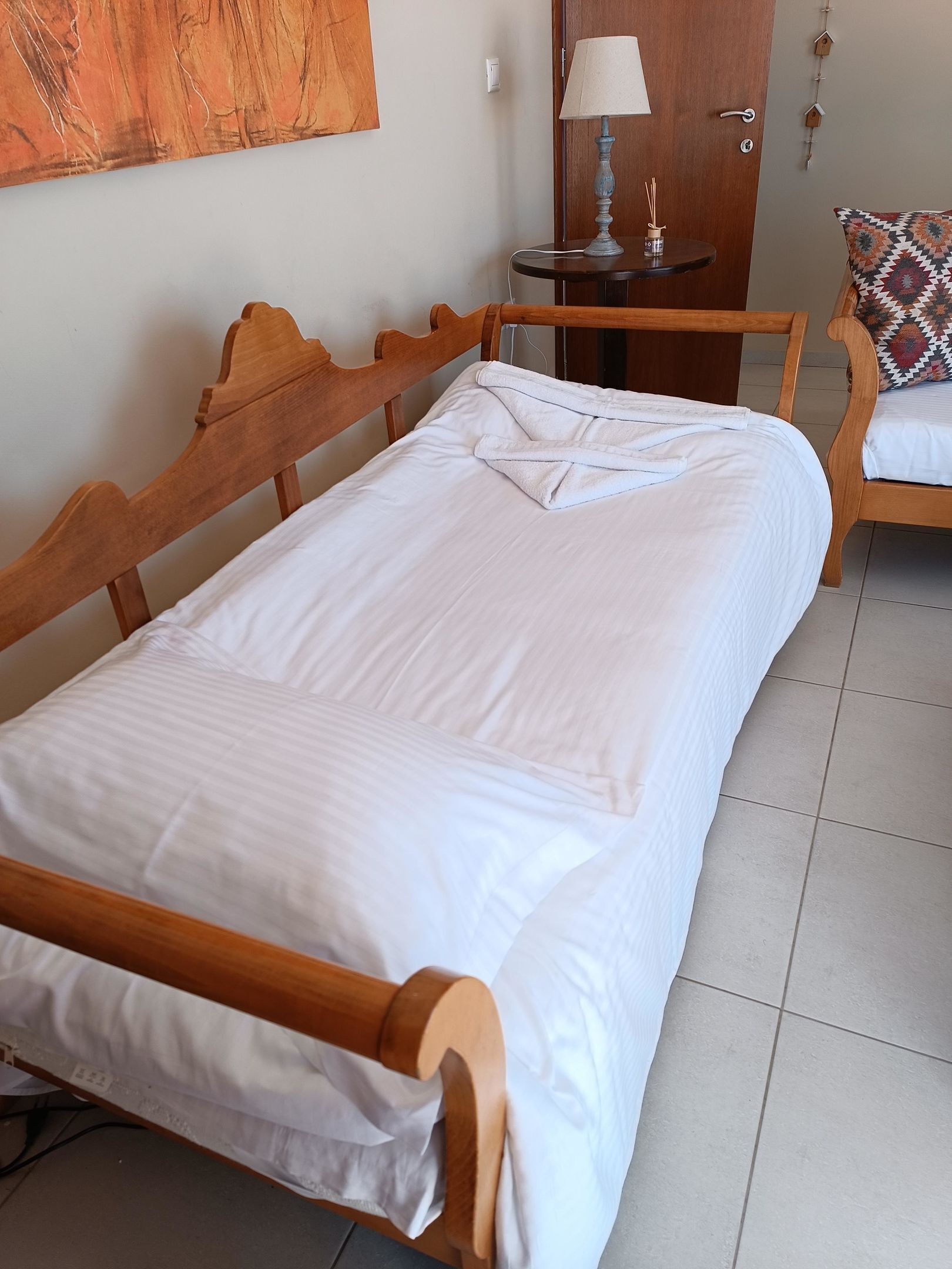 A neatly made bed with a wooden headboard, a small bedside table with a lamp, and a patterned pillow on a wooden chair.