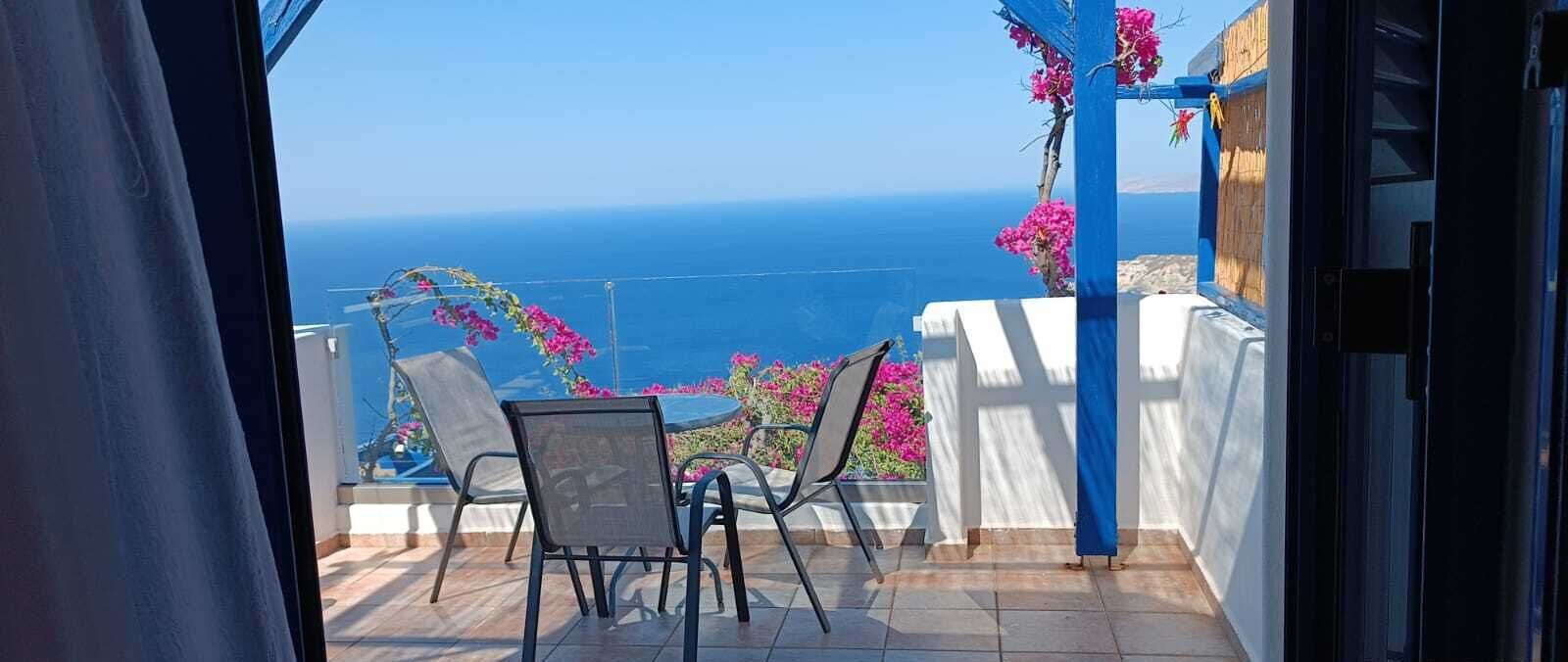 A balcony with a view of the sea, a table with chairs, and pink flowers.