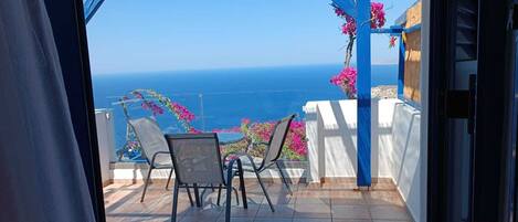 A balcony with a view of the sea, a table with chairs, and pink flowers.