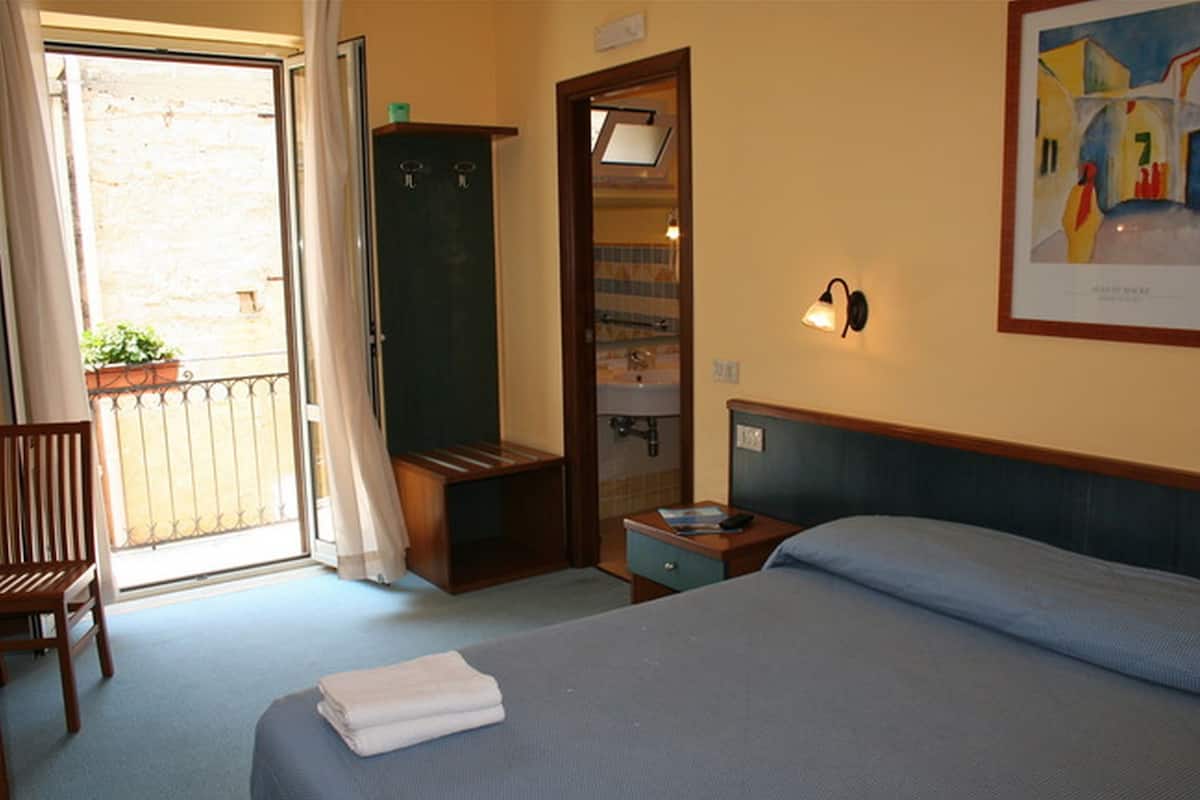 A hotel room with a bed, a chair, a wardrobe, a small table, and a painting on the wall.