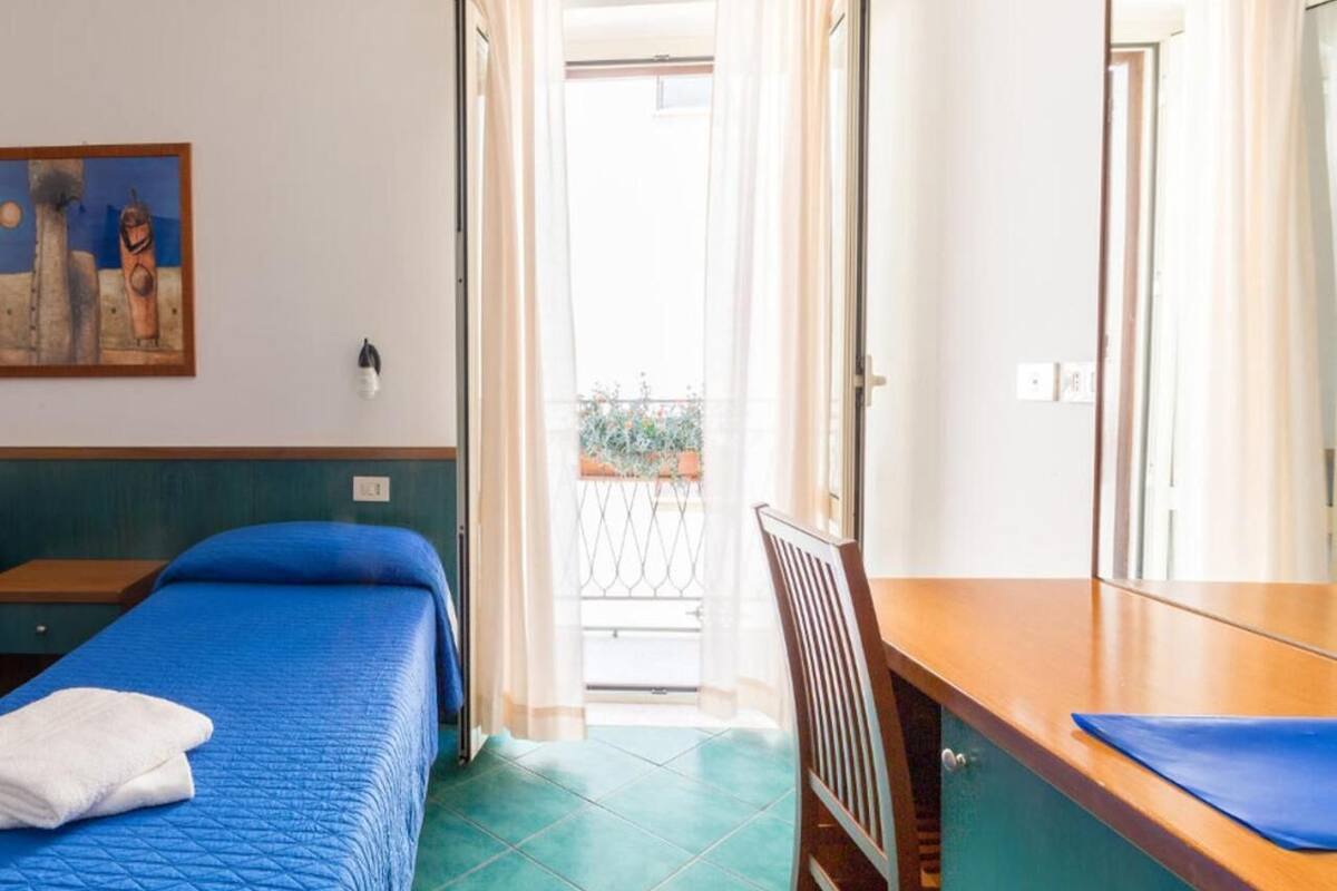 A hotel room with a bed, a desk, and a chair. There is a balcony with a view of the outside.