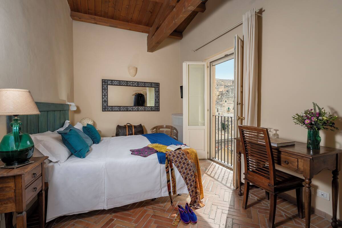 A bedroom with a bed, a desk, a chair, a lamp, and a window with a view of buildings.