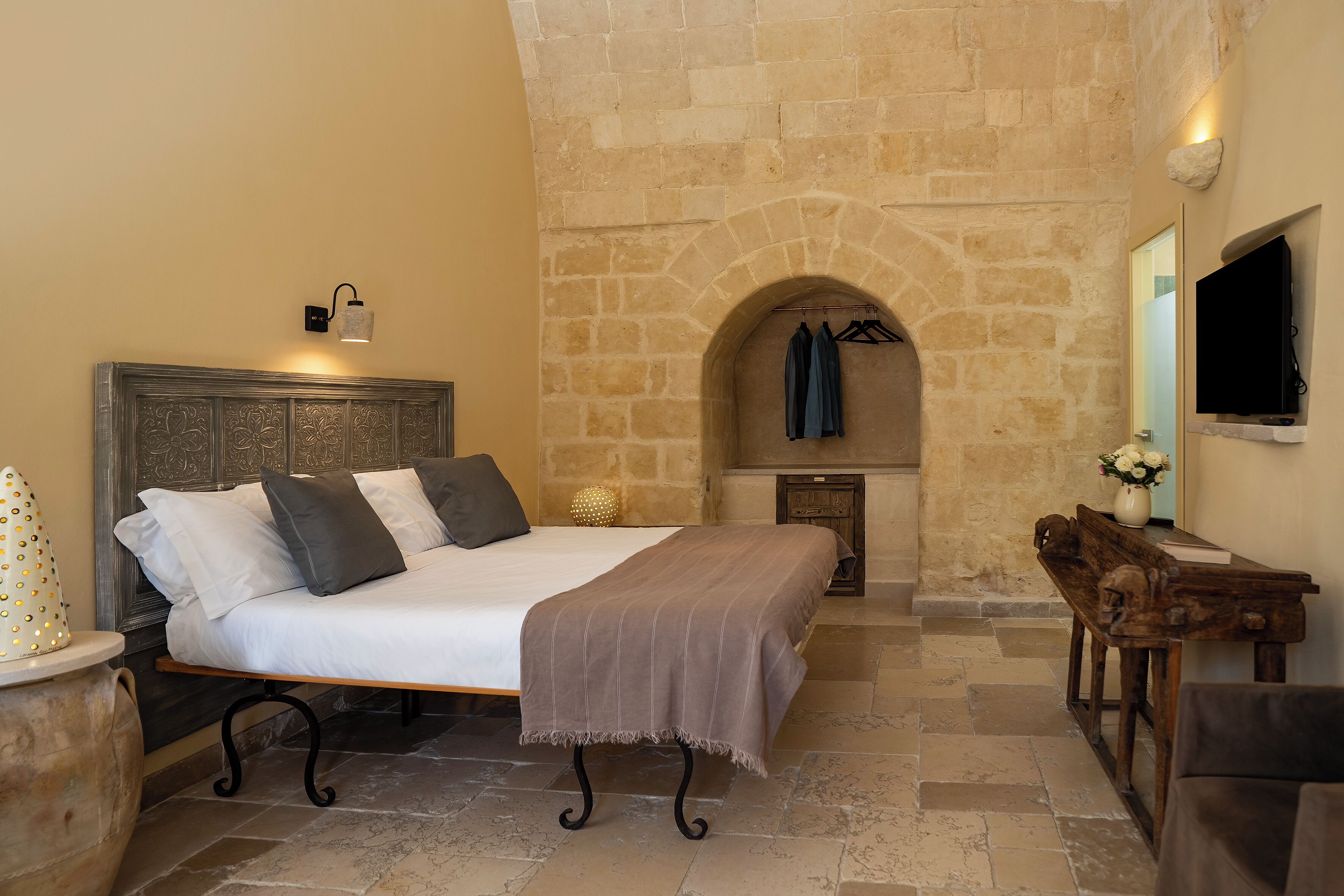 A bedroom with a stone wall, a bed with a headboard, a bedside table, a wooden bench, and a TV mounted on the wall.