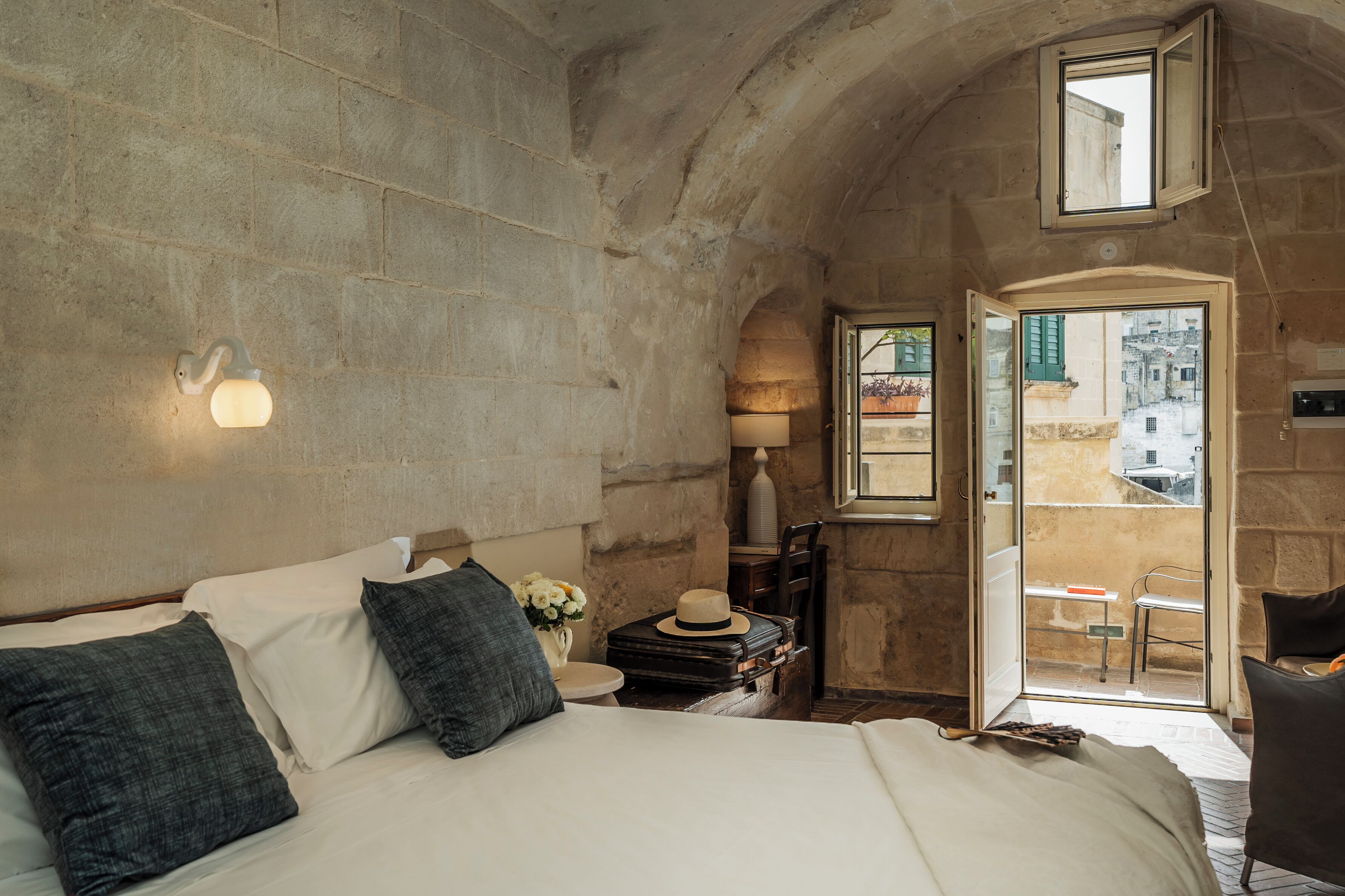 A bedroom with a stone wall, a bed with pillows, a nightstand with a lamp, and a suitcase.