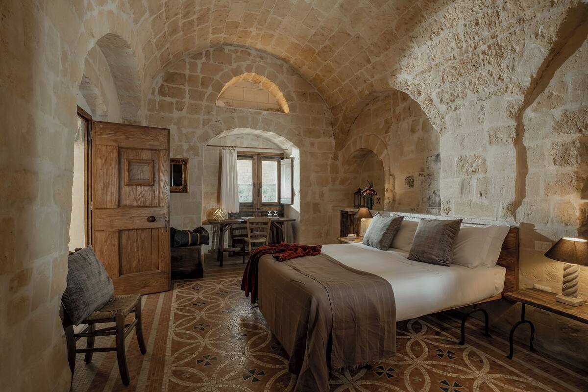 A rustic stone-walled room with a bed, a desk, and a chair.