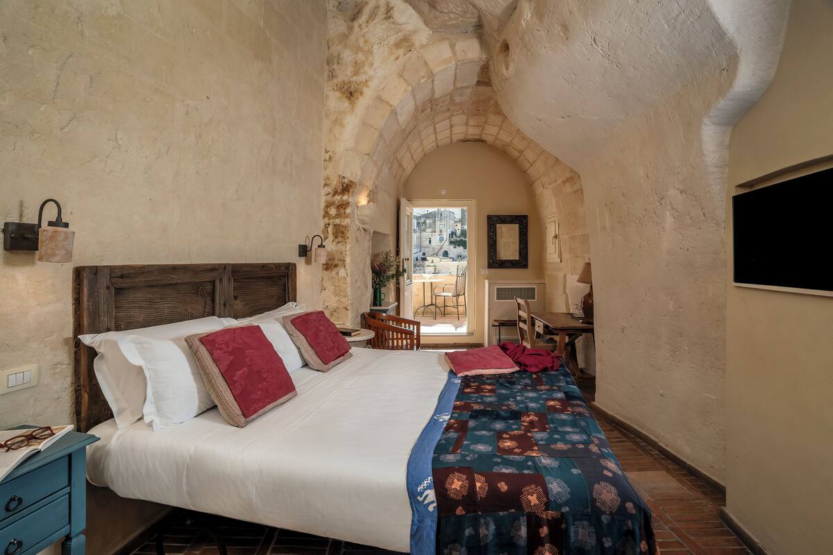 A bedroom with a large bed, a wooden headboard, a blue and red bedspread, and a stone archway leading to a balcony.