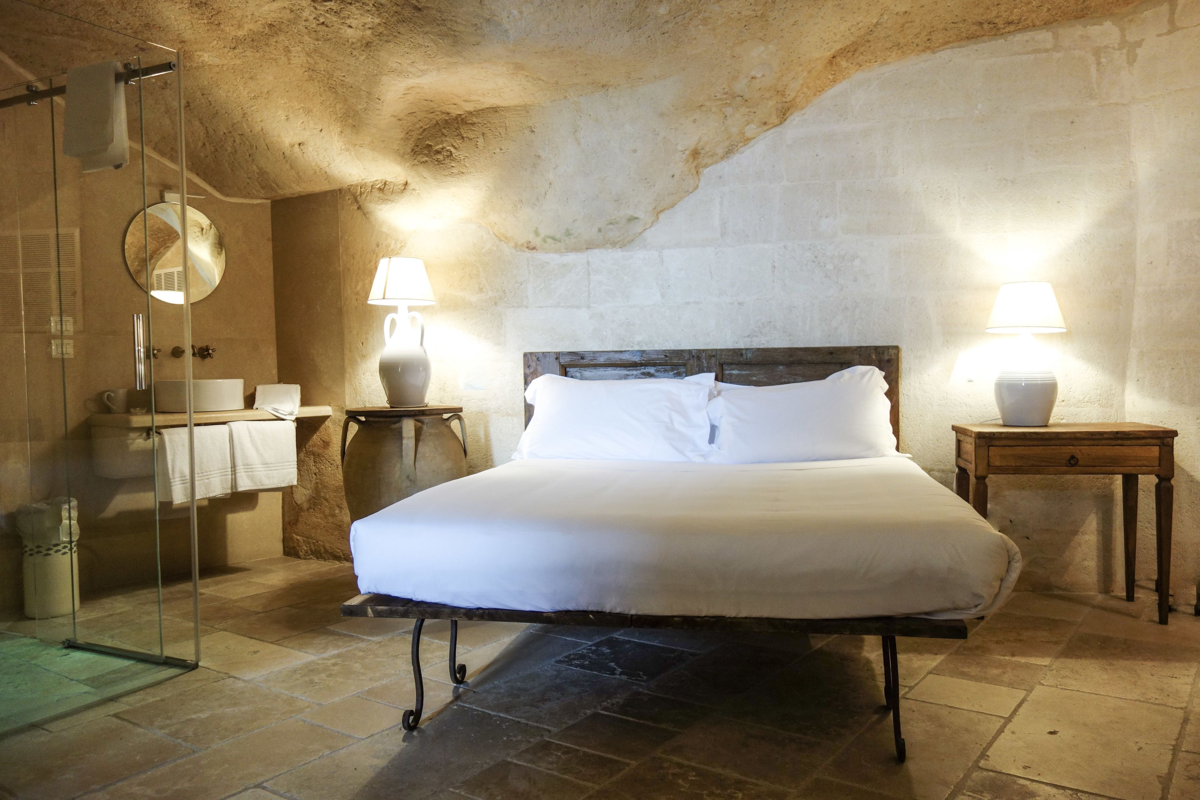 A bedroom with a large bed, a wooden headboard, a round mirror, a bedside table with a lamp, and a stone wall.