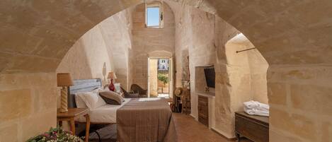 A rustic bedroom with a stone archway, a wooden bed, a TV, and a bathroom with a bathtub.