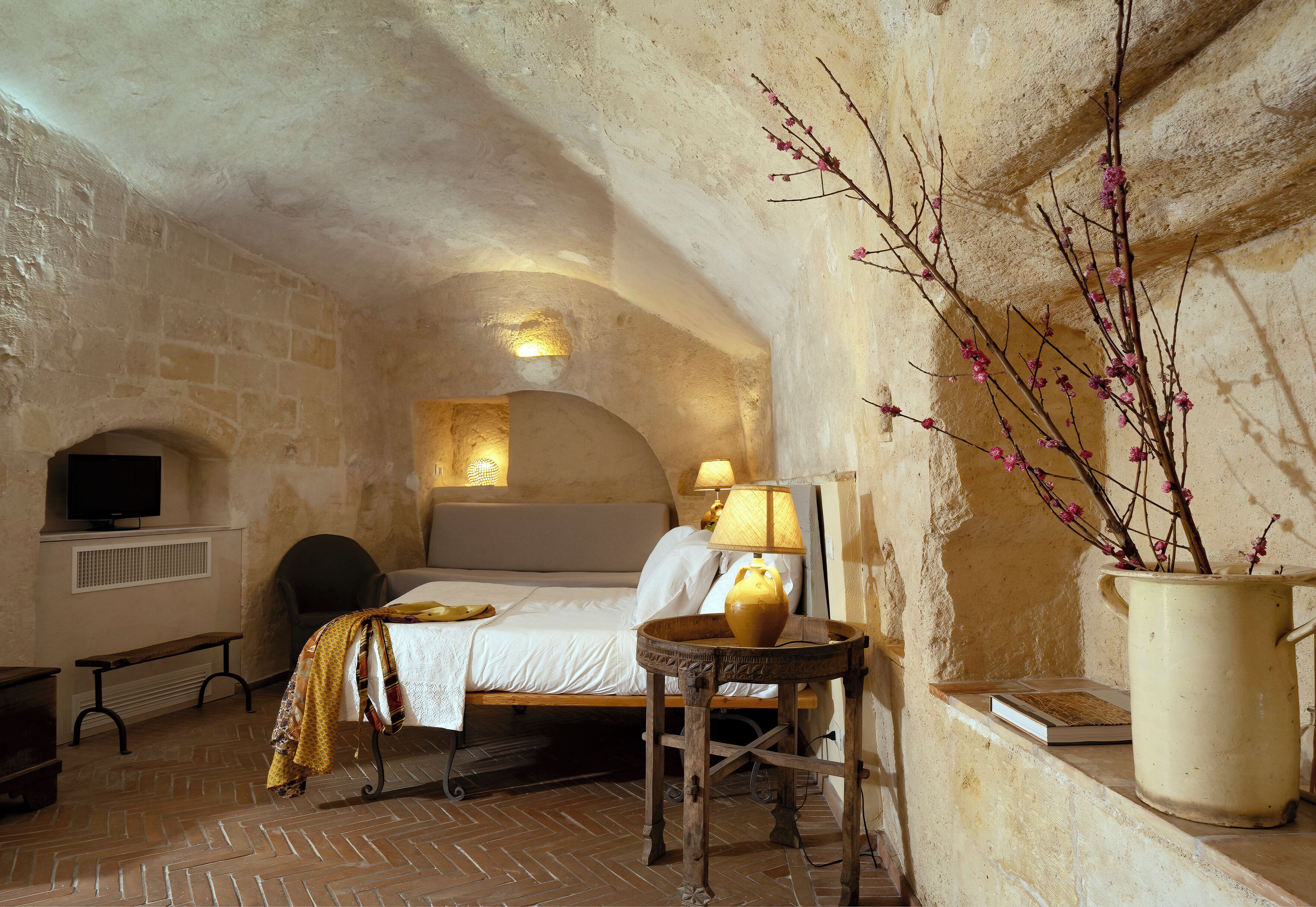 A cozy bedroom with a bed, bedside table, lamp, and a vase with branches.