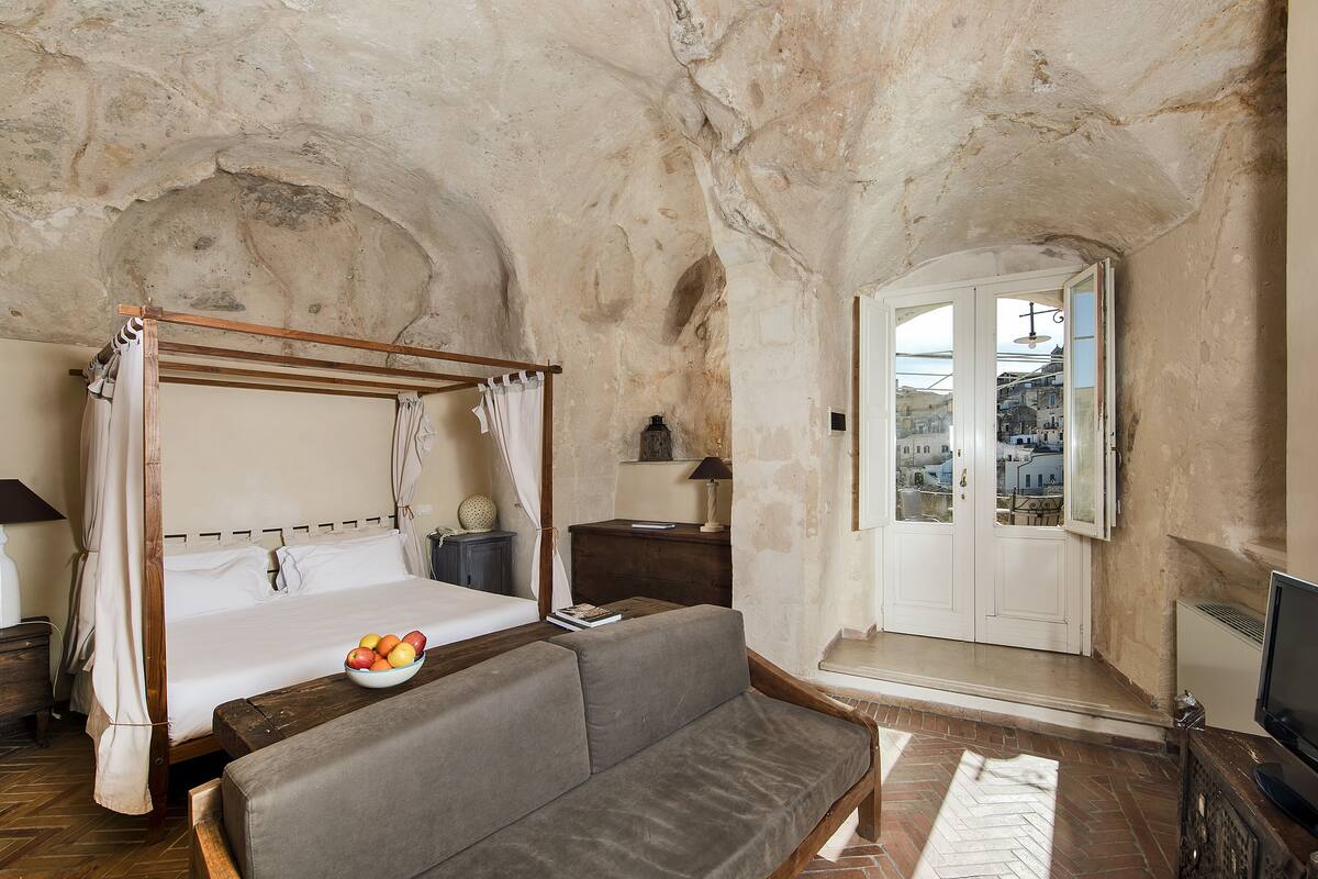 A bedroom with a four-poster bed, a sofa, a bowl of fruit, and a view of the city through a door.