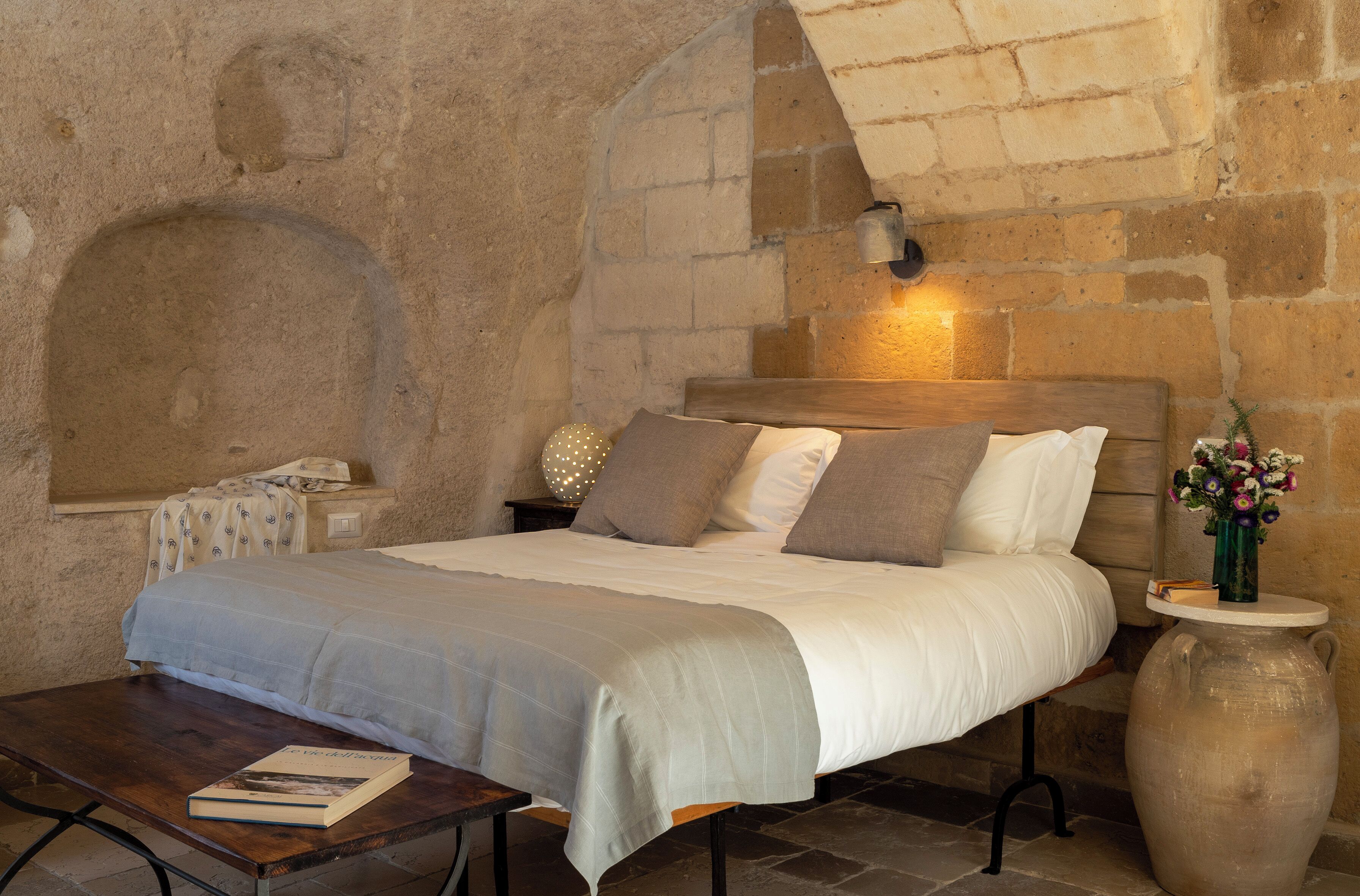 A bedroom with a stone wall, a wooden headboard, a bedside table with a lamp, and a large vase with flowers.