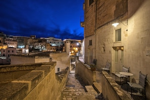 A narrow alleyway with stone walls, a small outdoor seating area with tables and chairs, and buildings with balconies and windows in the background.