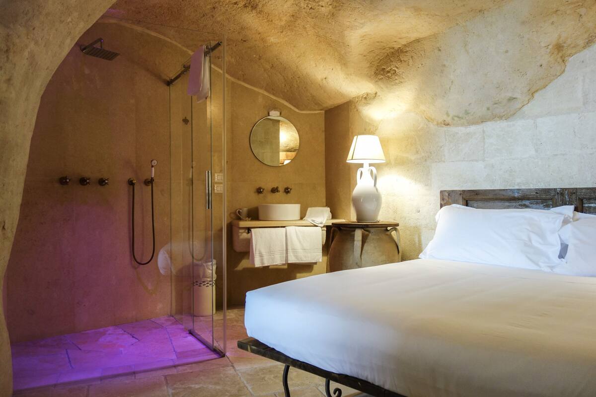 A cozy room with a bed, a bedside lamp, a round mirror, a sink, and a shower.