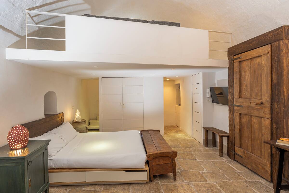 A bedroom with a loft bed, a wooden bench, a stone floor, a wooden wardrobe, and a small table with a book.