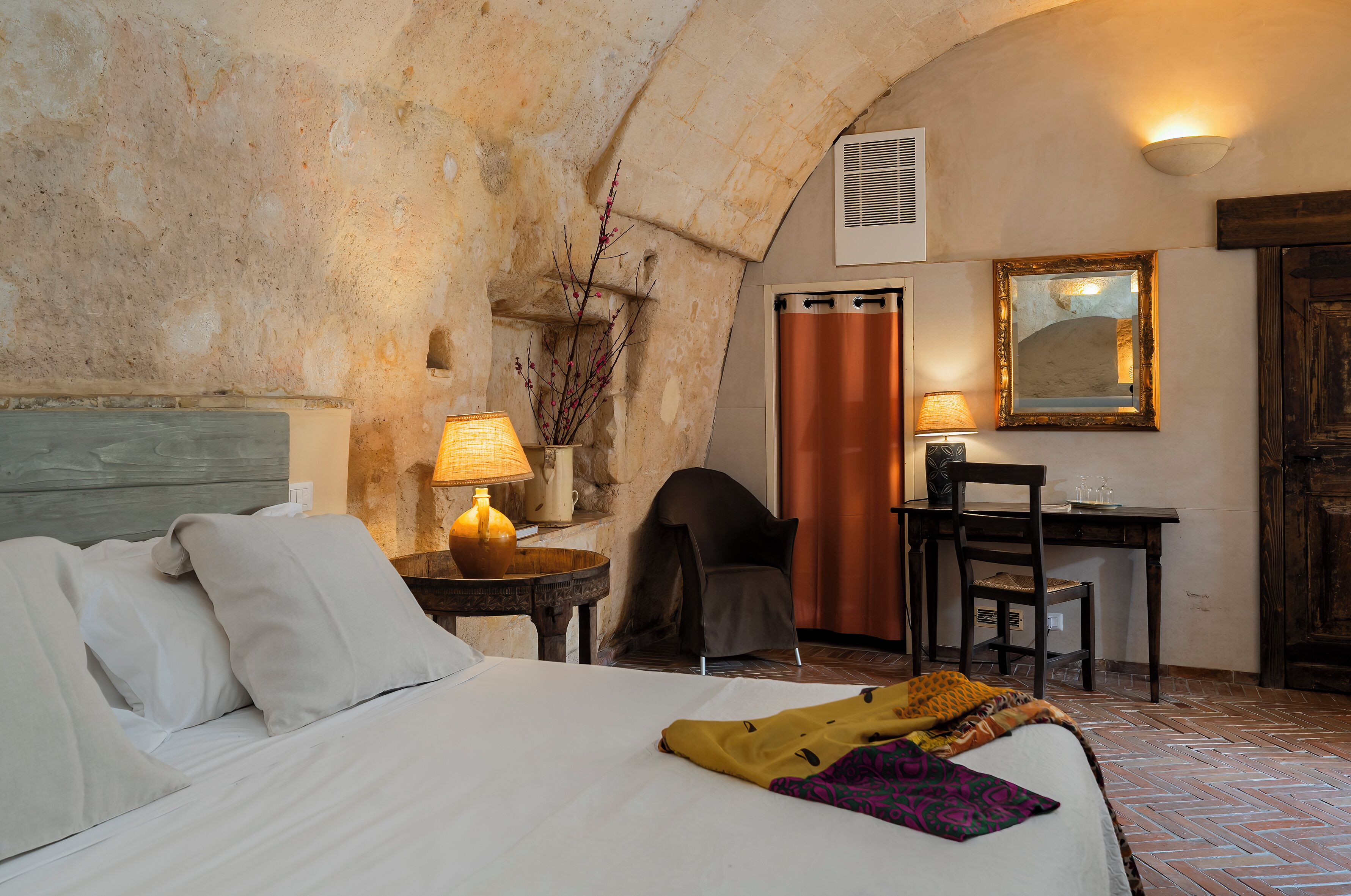 A hotel room with a bed, a desk, a chair, a lamp, and a mirror.