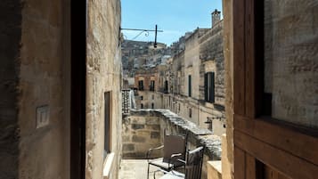 A narrow stone corridor leading to a balcony with a table and chairs, overlooking a historic town with stone buildings and a clear sky.