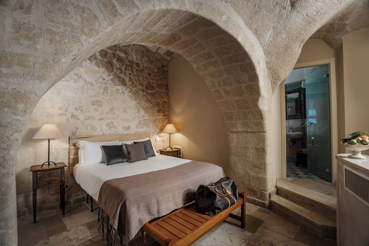 A bedroom with a stone archway, a bed with a white and brown bedspread, a wooden bench, a nightstand with a lamp, and a view of a bathroom through a glass door.