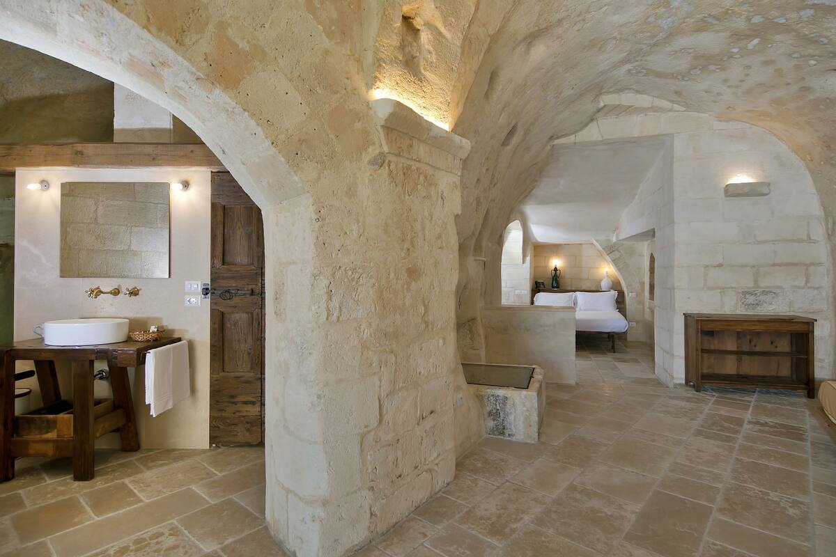 A rustic stone interior with a stone sink, wooden stool, and a mirror.