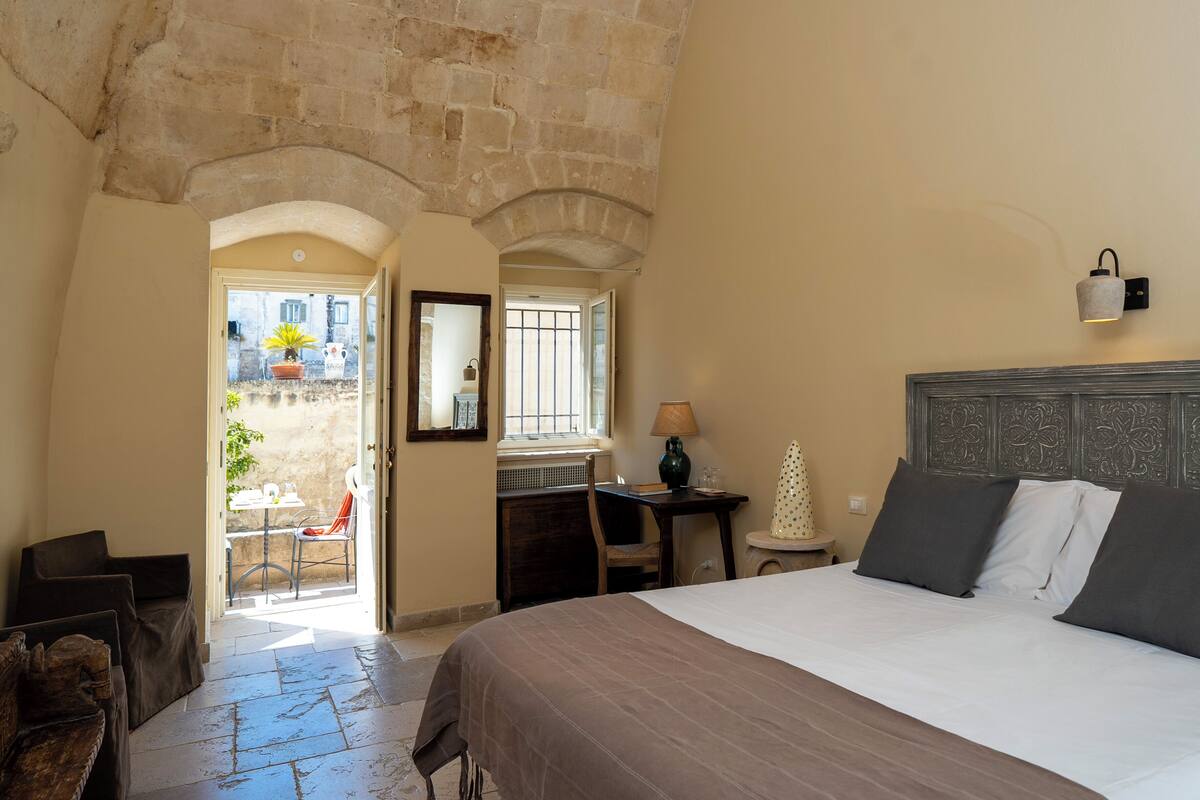 A bedroom with a bed, a desk, a chair, and a small table. There is a window with a view of the outside.
