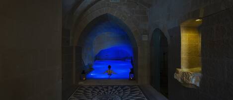 Un pasillo de piedra con una piscina iluminada en azul, suelo con diseño y paredes de piedra.