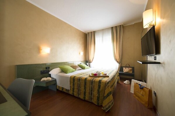 A hotel room with a bed, bedside table, lamp, TV, and a view of the outdoors. - Hotel Il Castelletto (Casarile)
