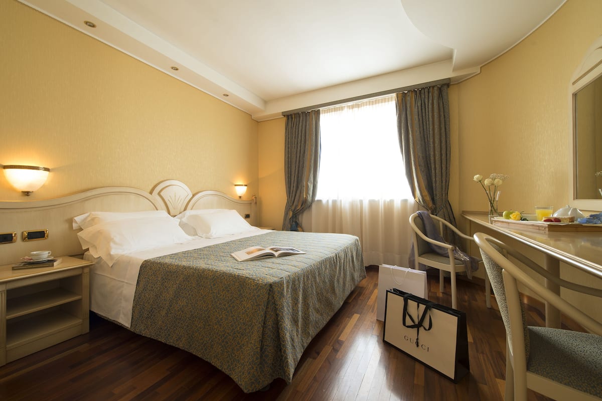 A hotel room with a bed, bedside table, lamp, chair, desk, and a window with curtains.