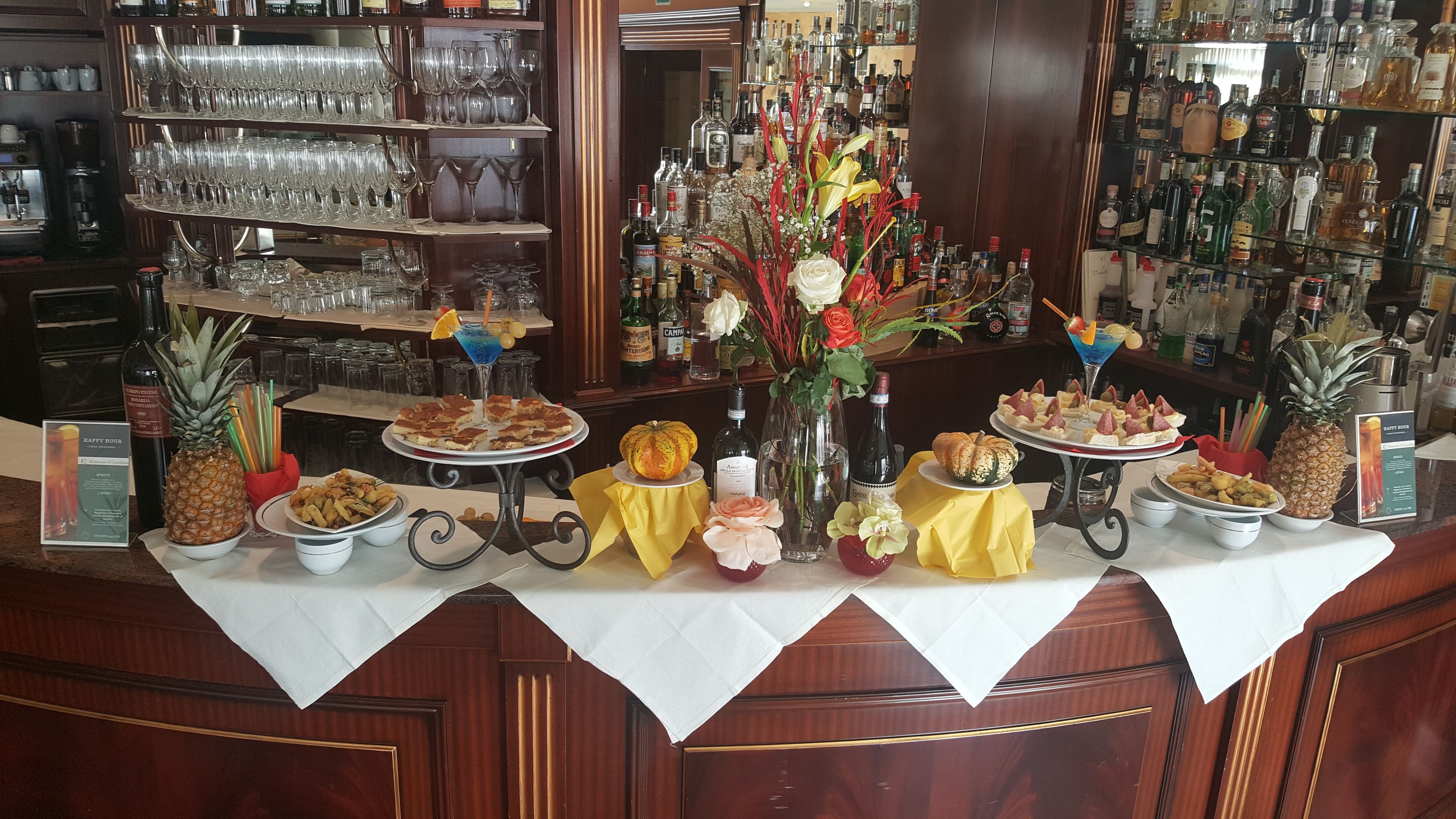 Angolo bar con numerose bottiglie di liquori, diversi bicchieri e una selezione di cibi e bevande sul bancone.