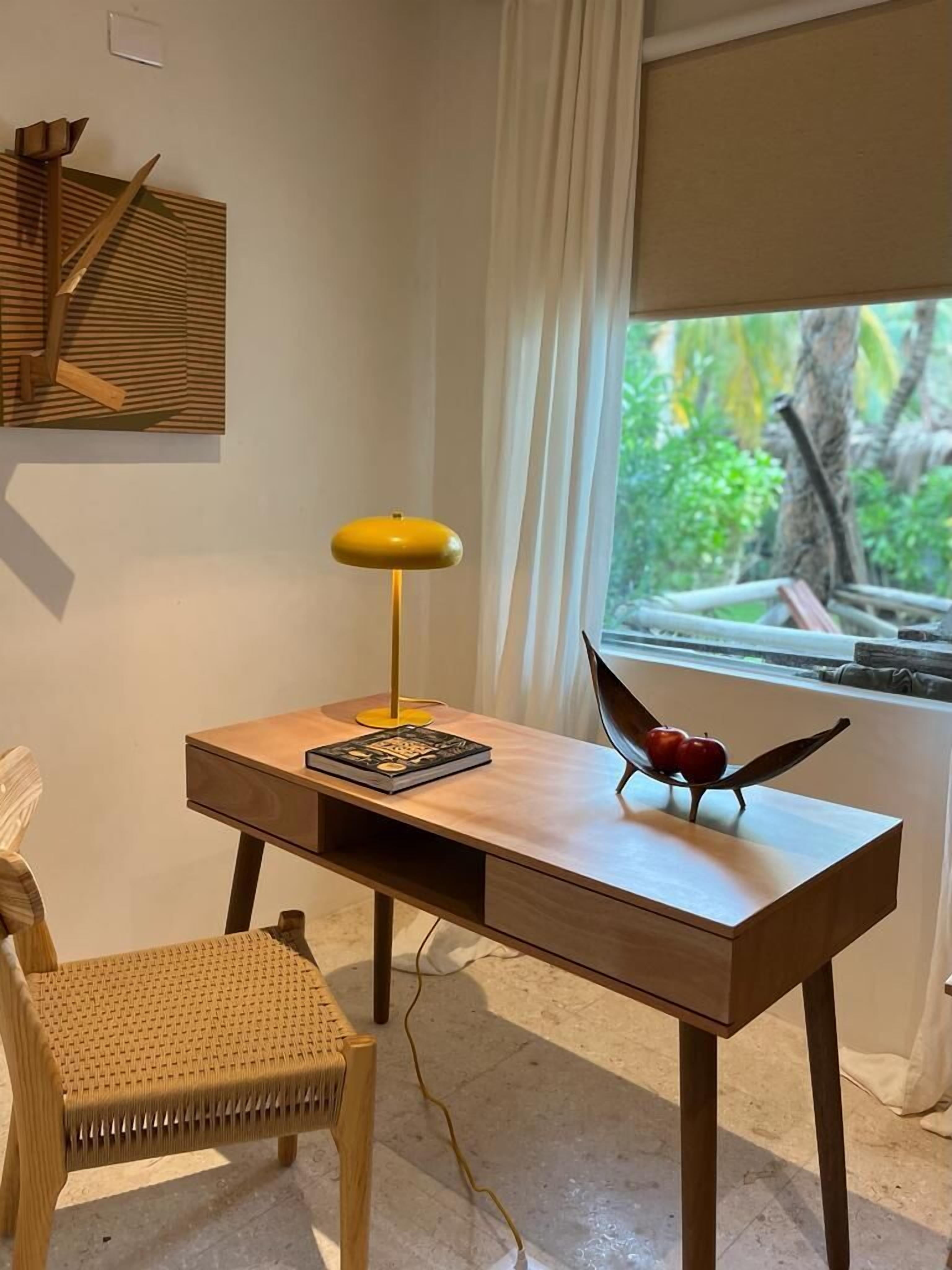 Una habitación moderna con un escritorio de madera, una lámpara de pie amarilla, una silla de mimbre y vistas a la vegetación a través de una ventana.
