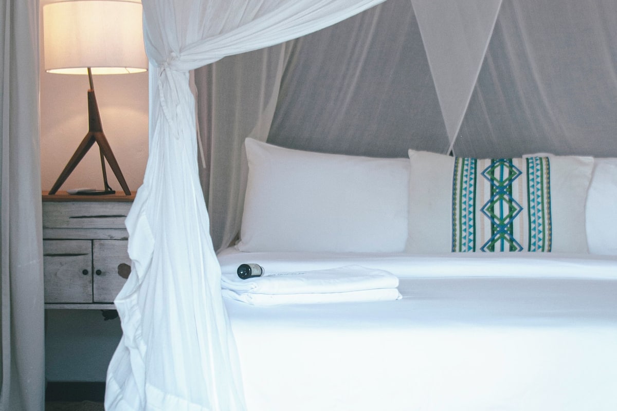 A bed with a canopy, a bedside table with a lamp, and a patterned pillow.