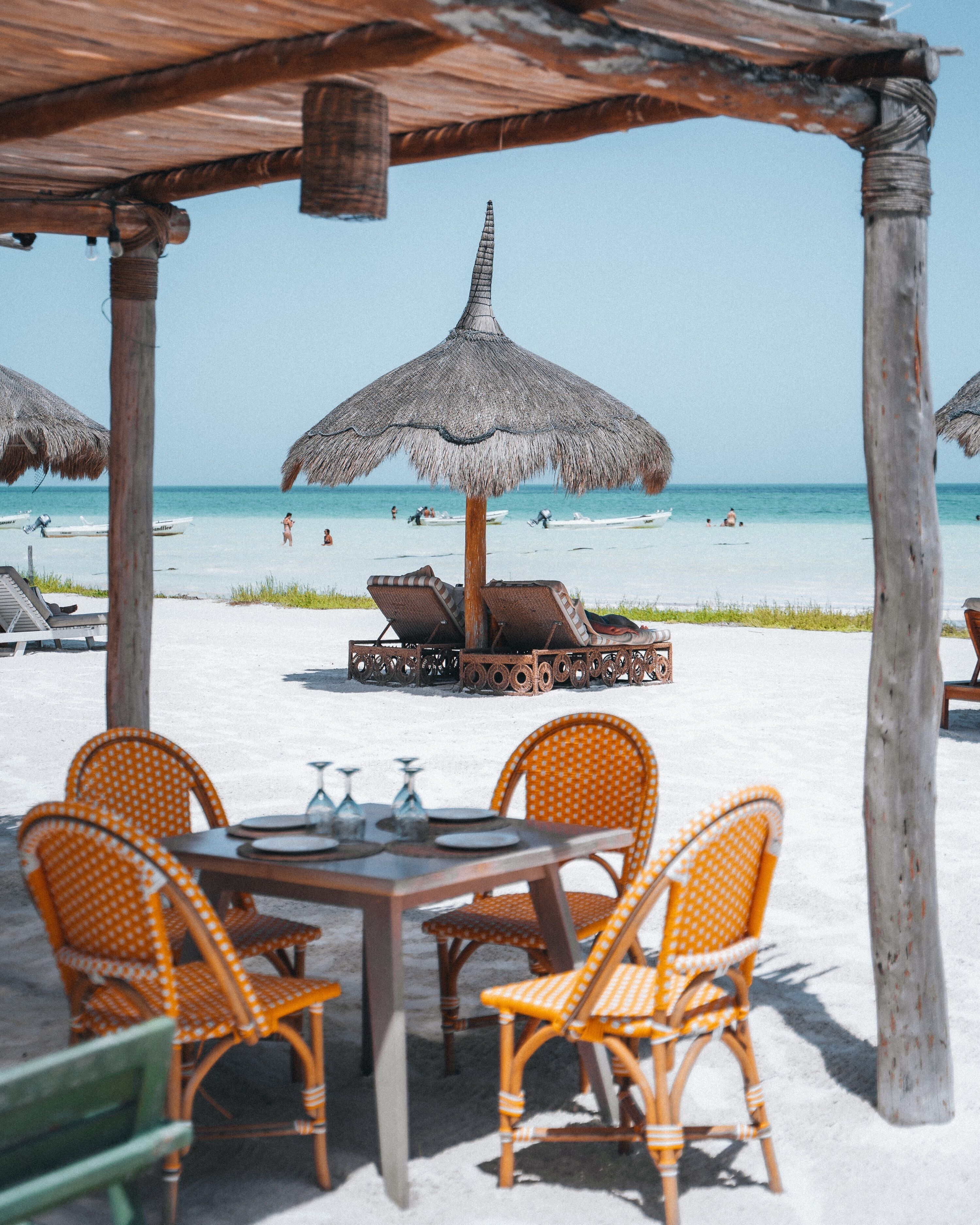 Un área de comedor frente a la playa con sillas de mimbre y una mesa, una sombrilla de paja, y vistas a la playa y al océano.