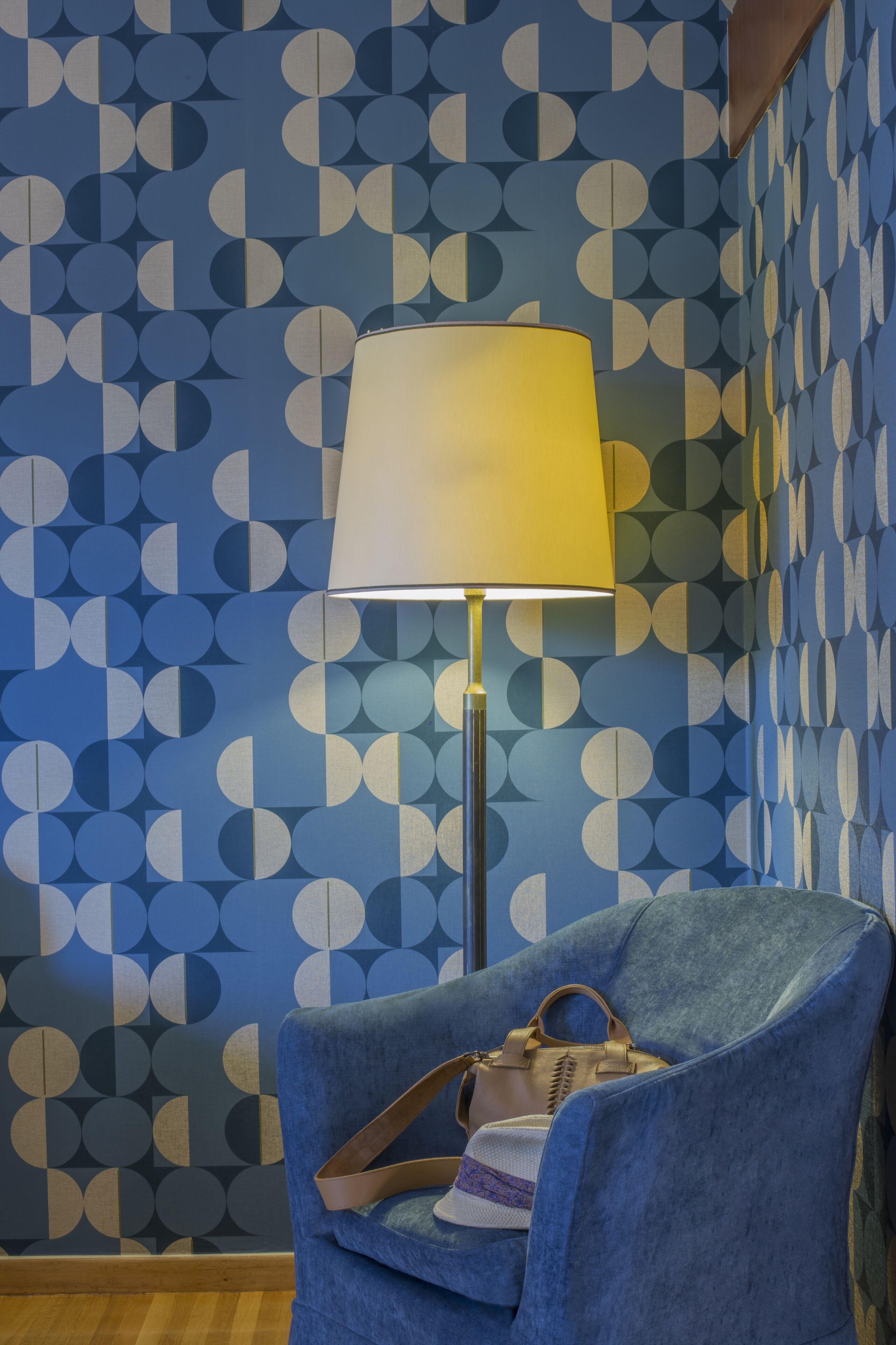 A room with a patterned wallpaper, a blue armchair, a lamp, and a handbag on the chair.