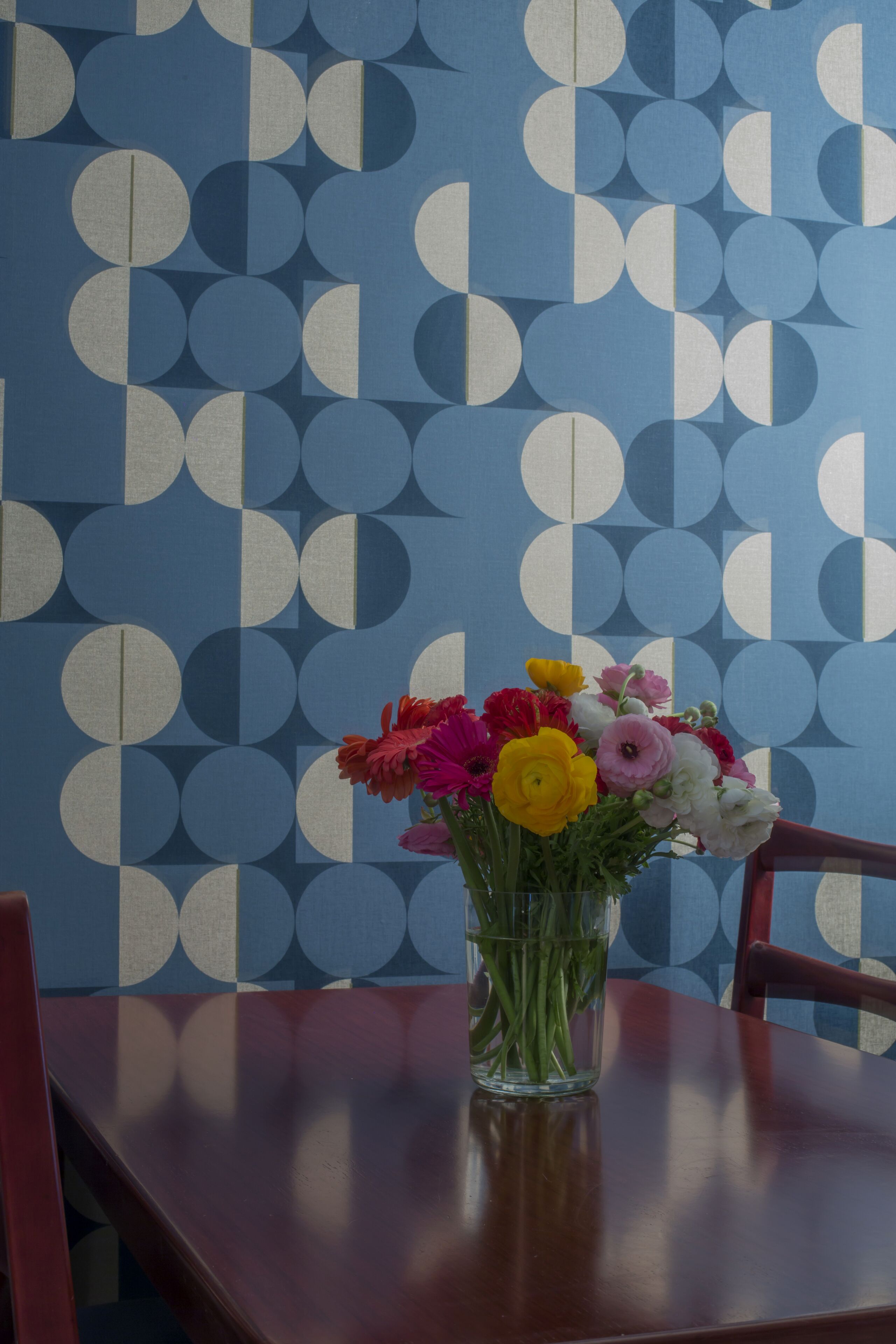 Una mesa con un jarrón de flores y un papel tapiz estampado al fondo.