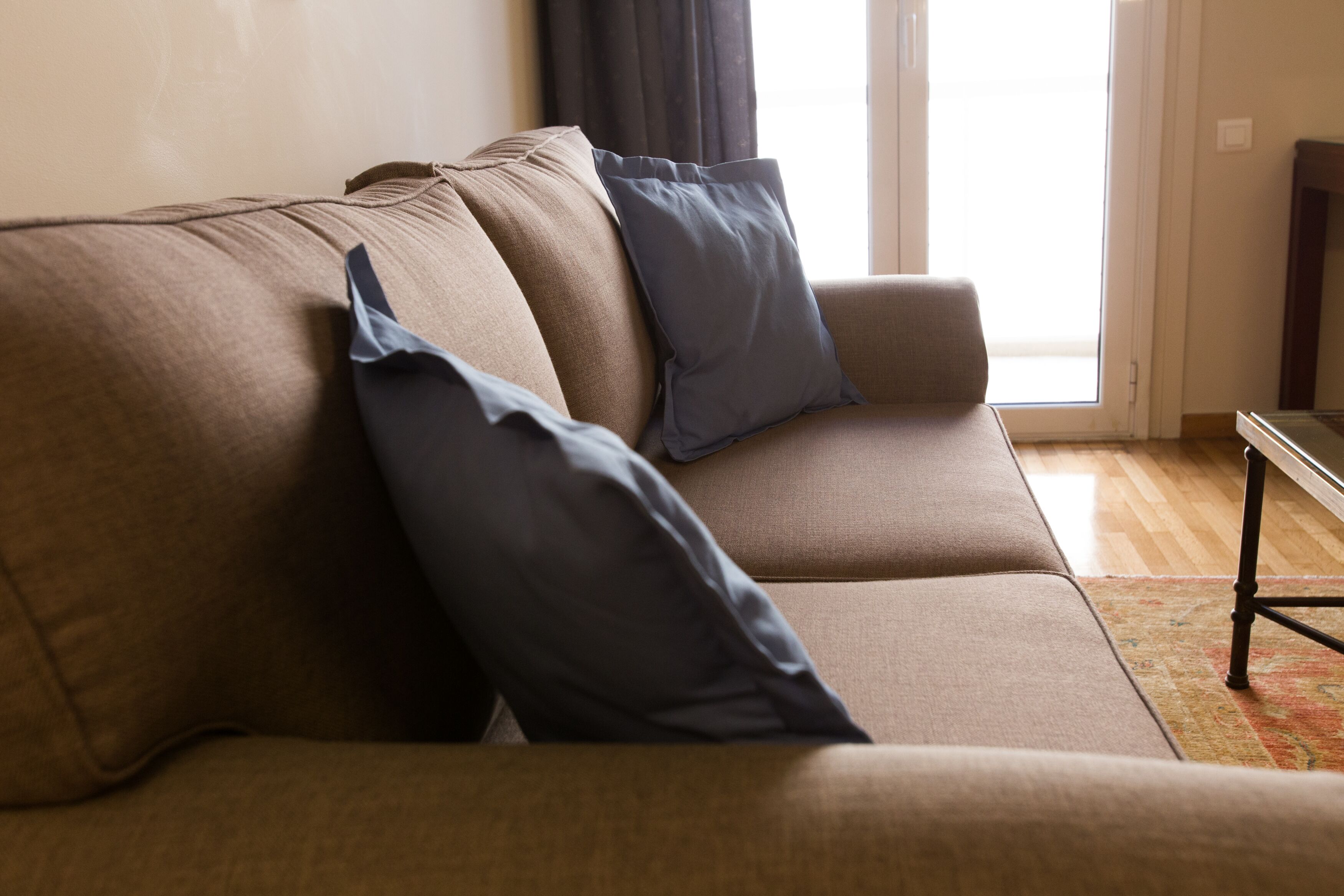 A beige sofa with two dark blue pillows, a wooden floor, and a glass door leading outside.