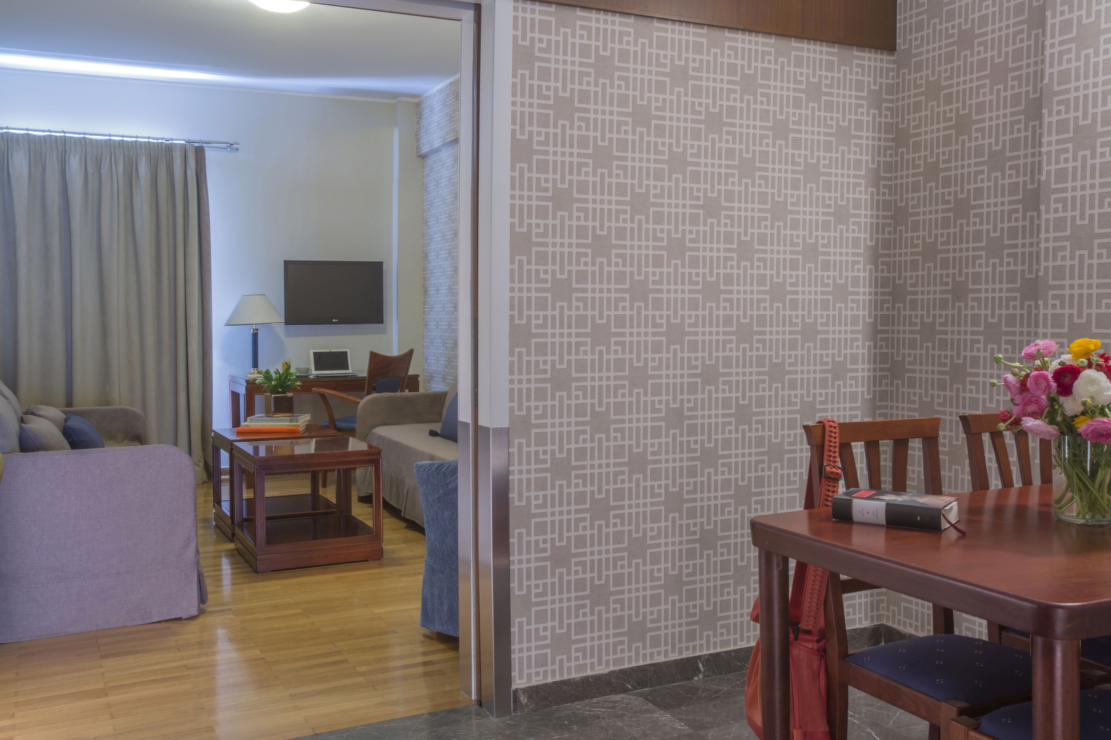 Une chambre d’hôtel comprenant un coin salon, une table à manger avec des chaises, un téléviseur fixé au mur et un papier peint à motifs.