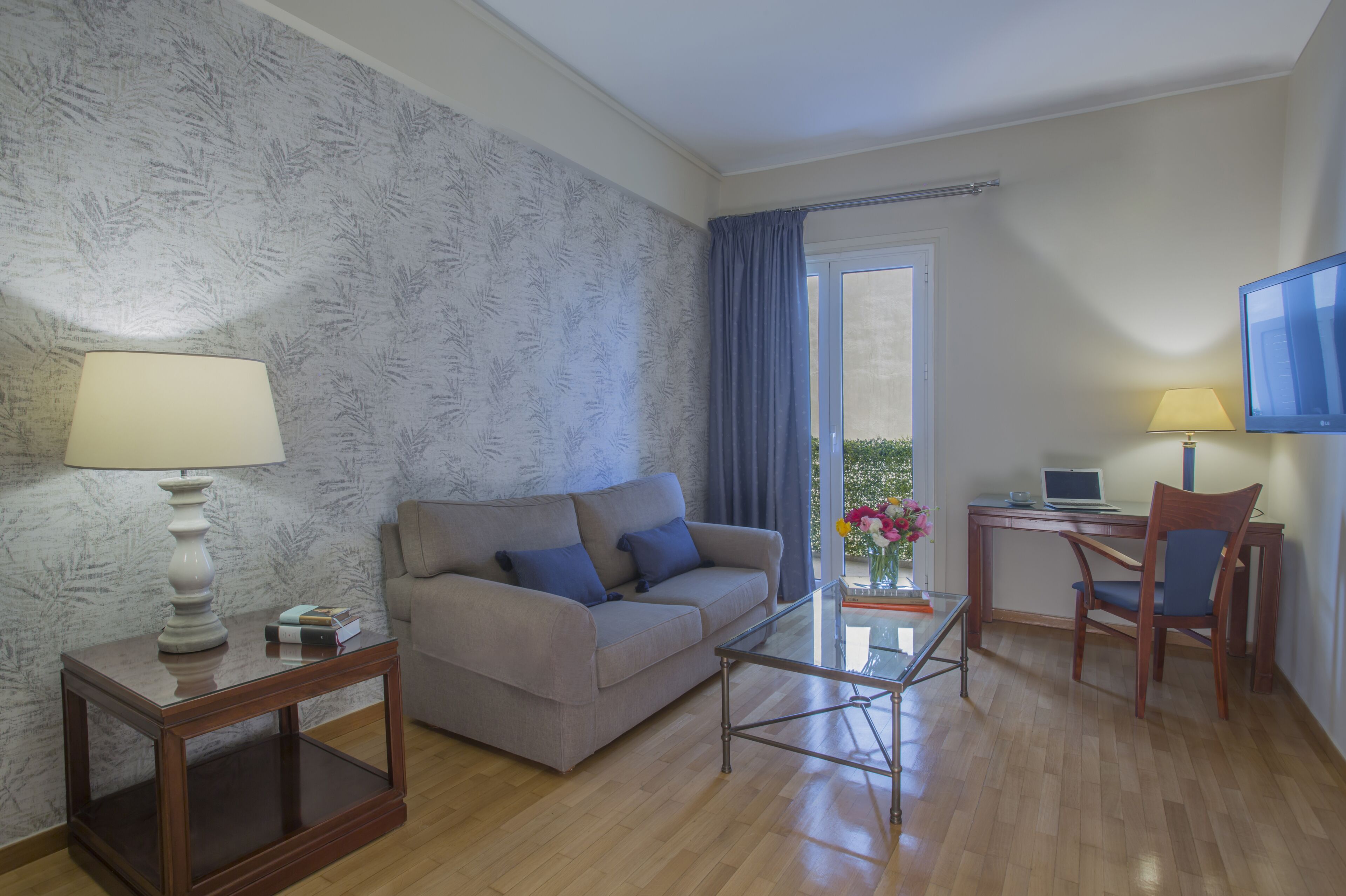 Una sala de estar con sofá, mesa de centro y escritorio con silla. Hay un televisor en la pared y una ventana con cortinas.