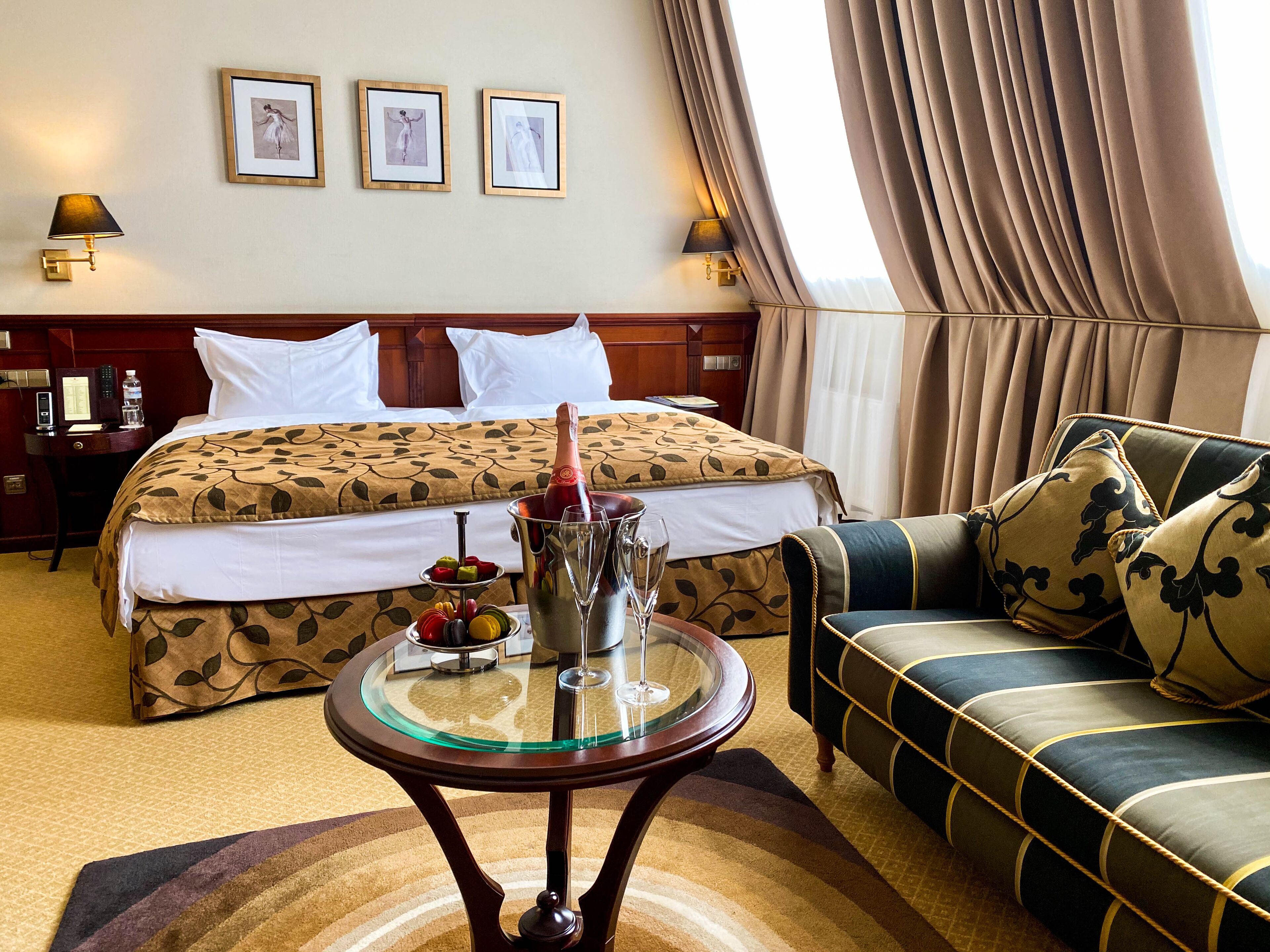 A hotel room with a bed, a sofa, a glass table with a bottle and glasses, and framed pictures on the wall.