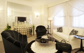 A modern living room with a black armchair, a white sofa with zebra-patterned pillows, a glass coffee table, and a black leather bag on the floor.