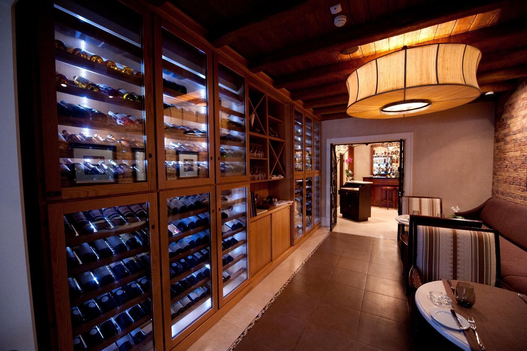 A wine cellar with wooden racks and glass doors, a bar area with stools, and a dining table set for two.