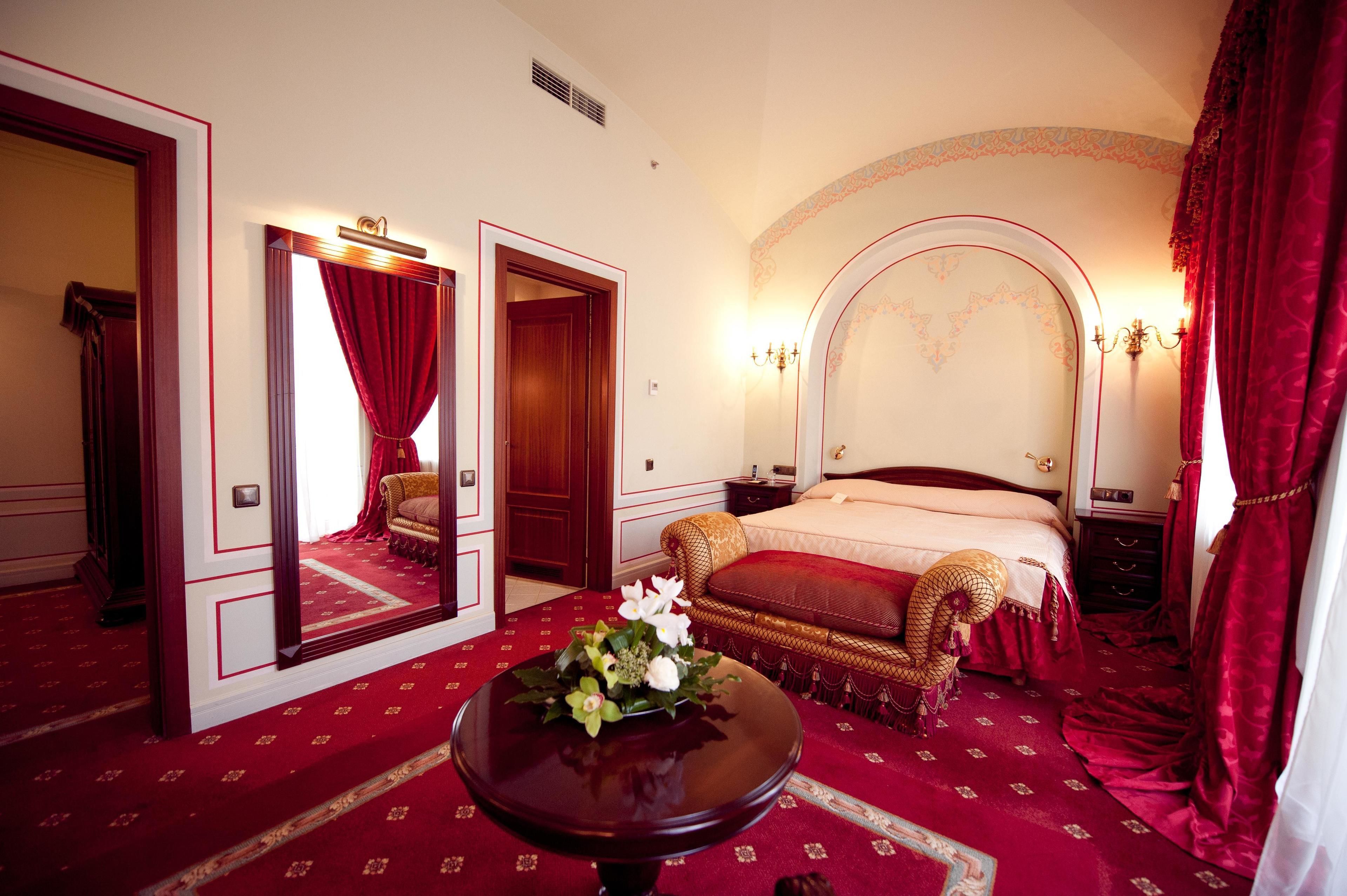 A hotel room with a large bed, a sitting area with a chaise lounge, a small table with a vase of flowers, and a mirror on the wall.